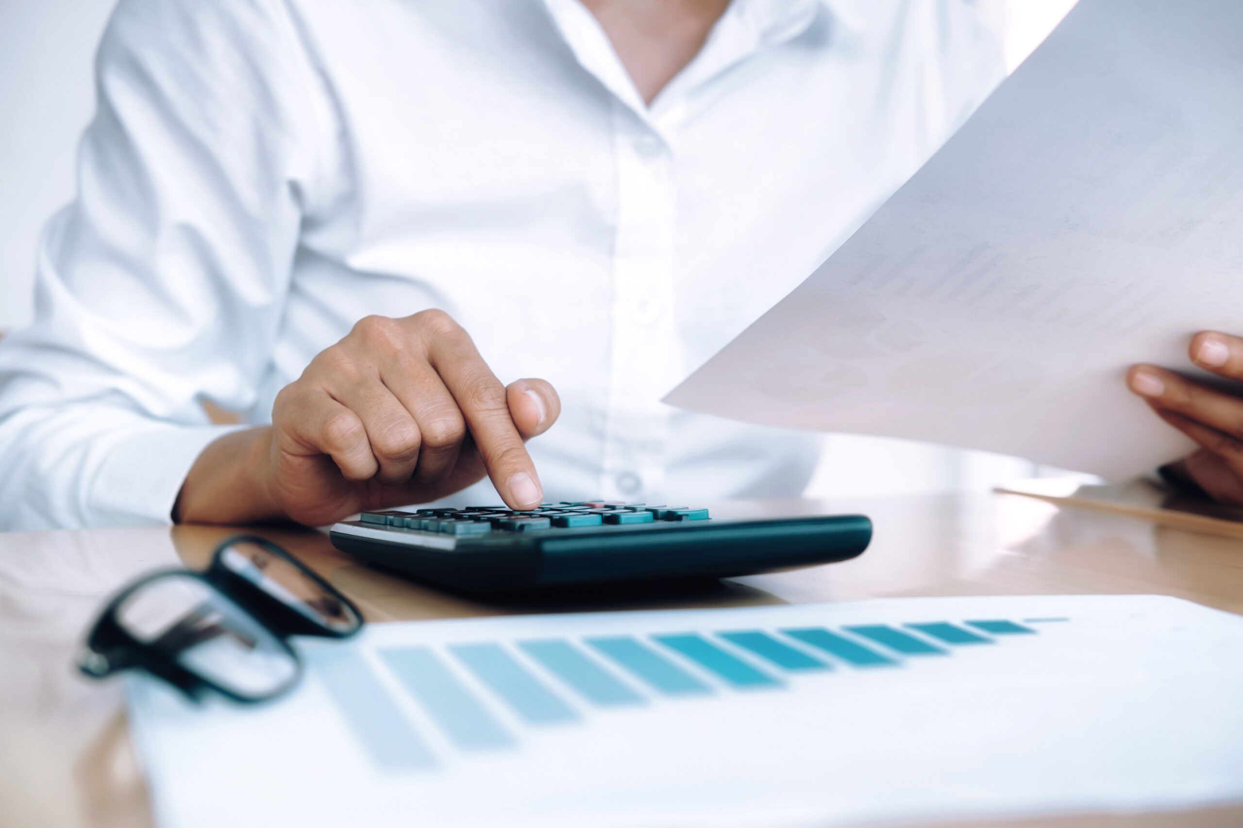 Pessoa utilizando uma calculadora enquanto analisa um documento financeiro sobre a mesa, com gráficos ao fundo, representando o cálculo e a apuração de tributos relacionados ao ICMS-ST em São Paulo.