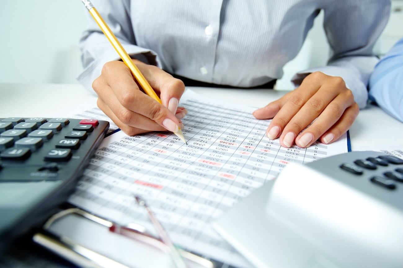 Imagem de mãos femininas escrevendo de lápis em planilha com calculadora ao lado fazendo fechamento contábil.
