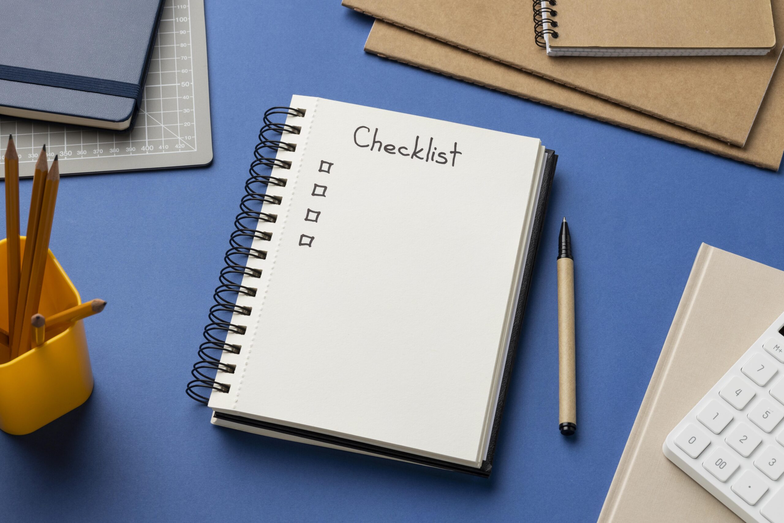 Caderno de checklist ao lado de lápis, caneta e teclado em uma mesa de trabalho.
