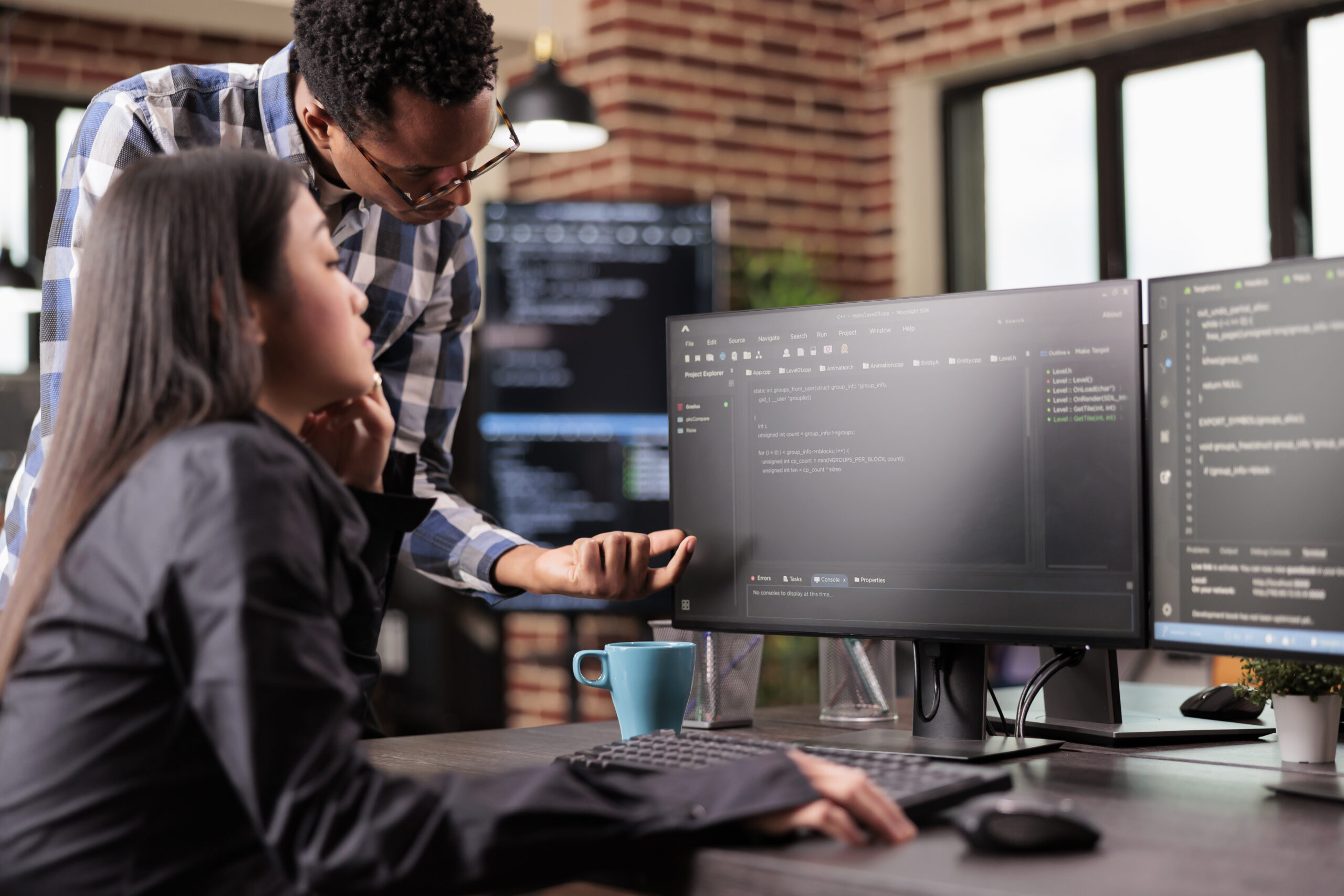 Dois profissionais analisam informações em monitores de computador em um escritório moderno, colaborando em atividades relacionadas a tecnologia, desenvolvimento de software e análise de dados.