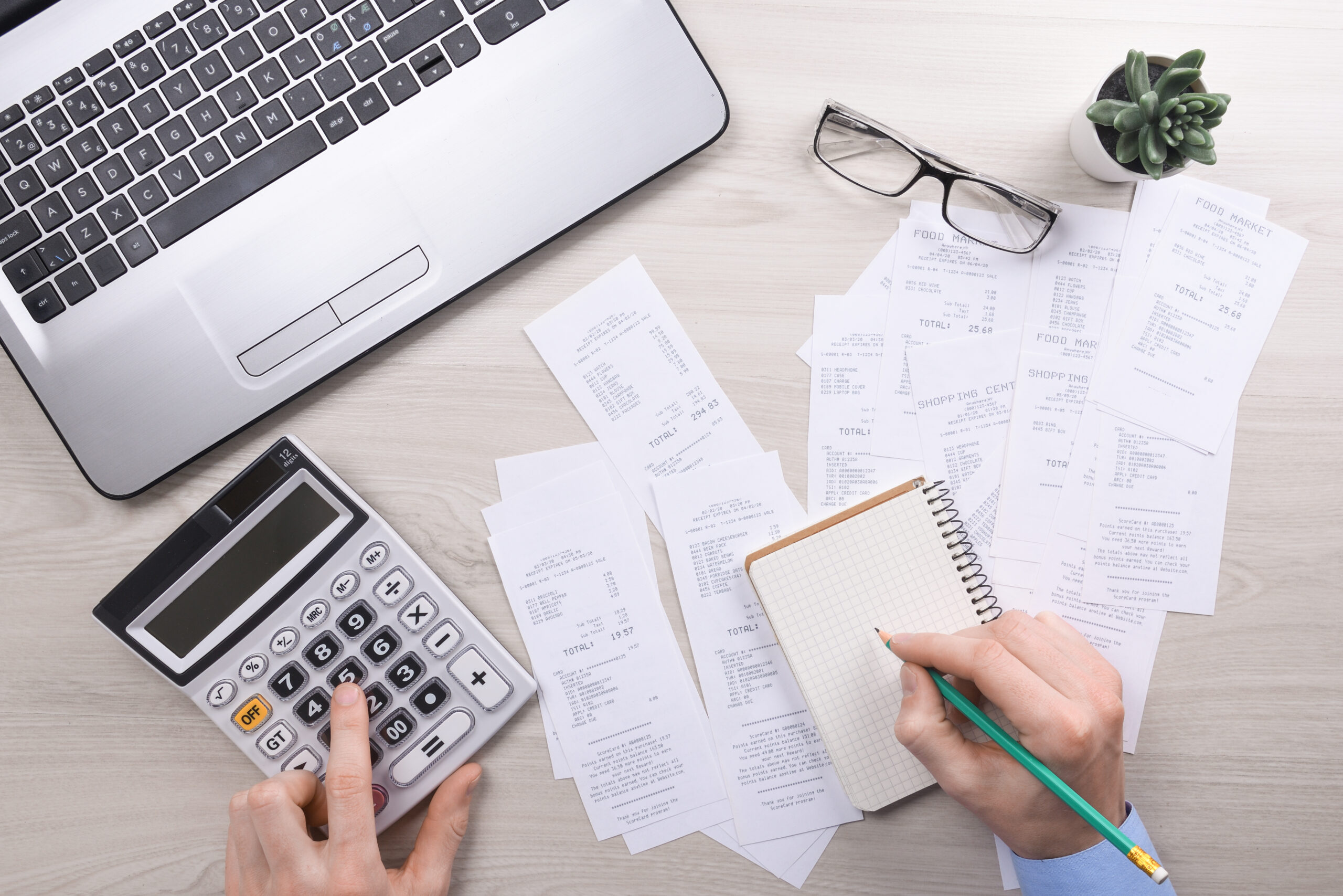 Pessoa utilizando uma calculadora enquanto registra anotações em um caderno, com diversos comprovantes fiscais espalhados sobre a mesa ao lado de um notebook.