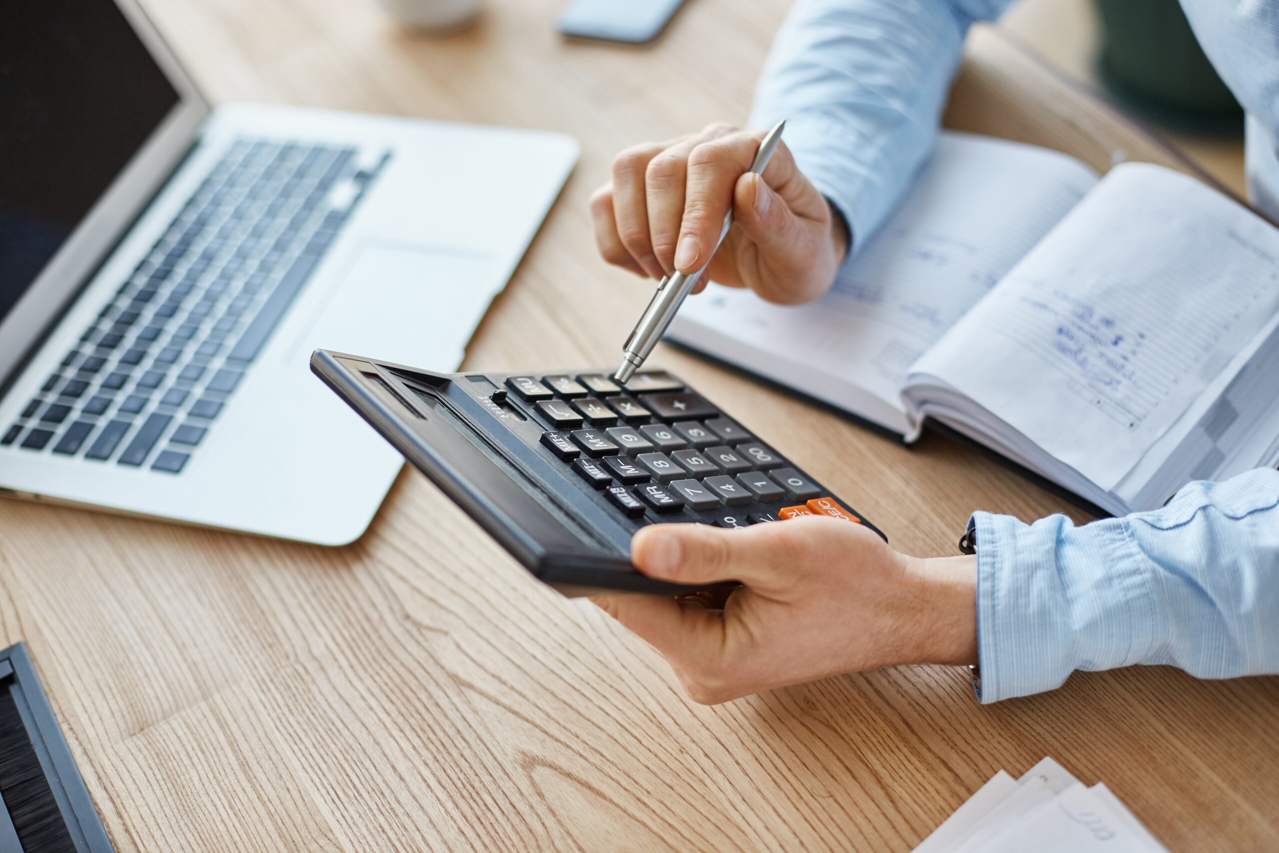 Pessoa utilizando uma calculadora de mesa enquanto faz cálculos financeiros em uma mesa de trabalho, com notebook aberto e caderno de anotações ao lado, representando análise de custos, precificação e planejamento tributário.