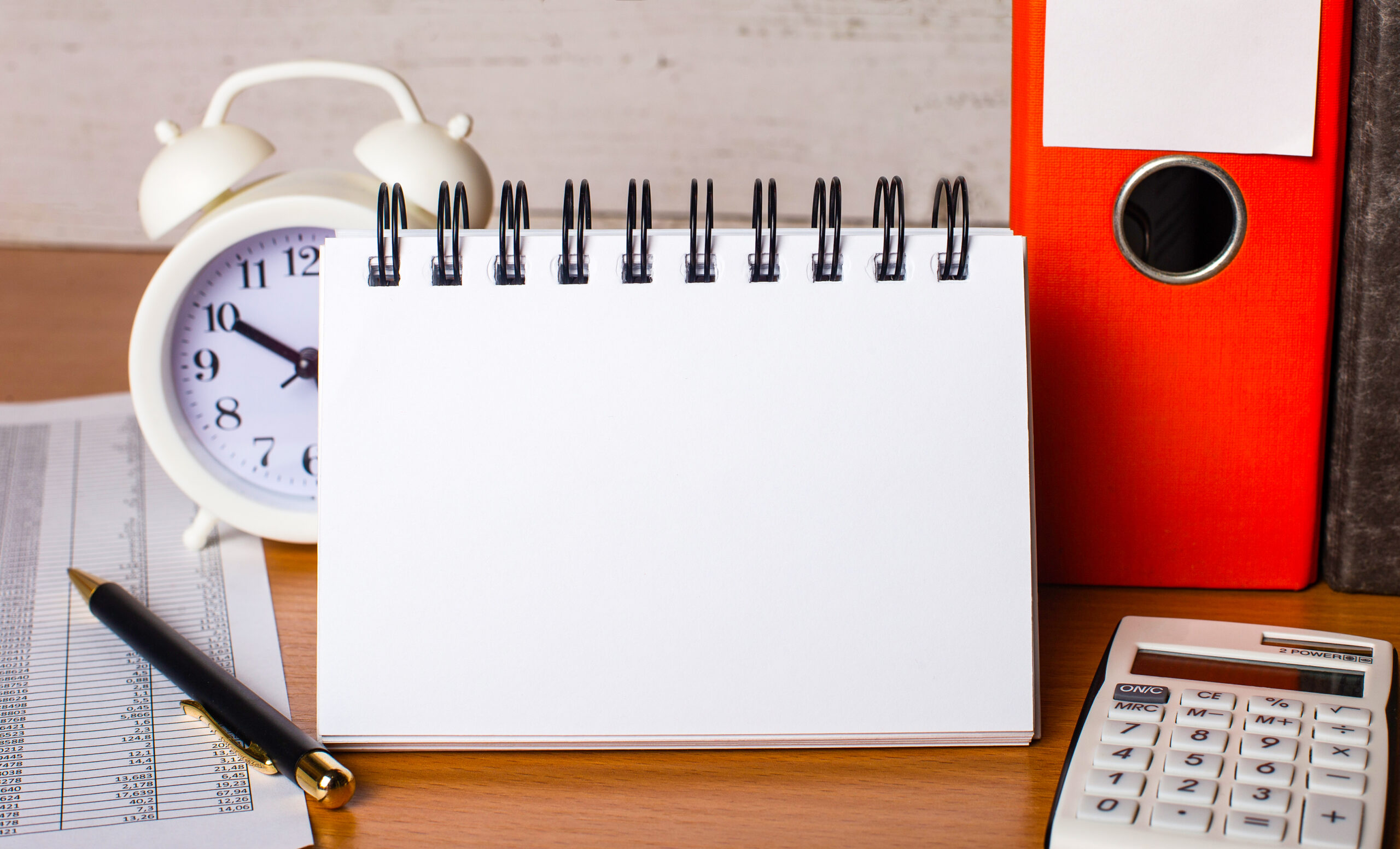 Mesa de escritório com calendário em branco, relógio despertador, calculadora, caneta, documentos fiscais, pasta de arquivos e bloco de anotações, representando a organização de prazos e obrigações fiscais.