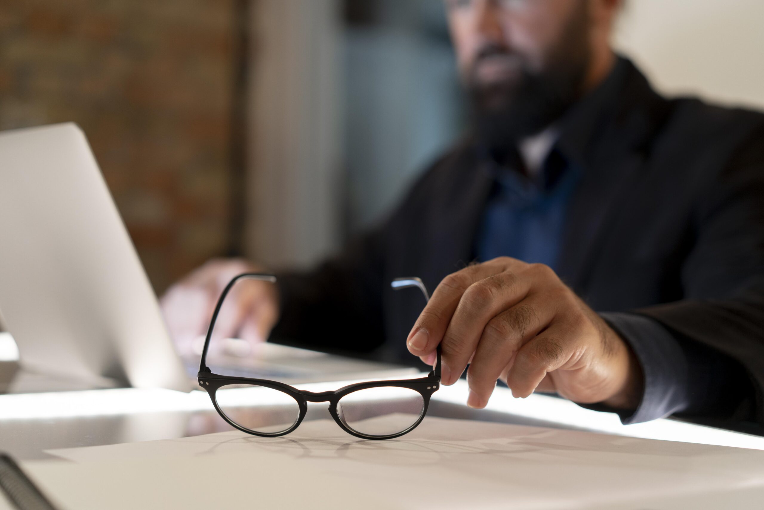 Profissional trabalhando no escritório enquanto analisa documentos ao lado de um notebook e óculos.