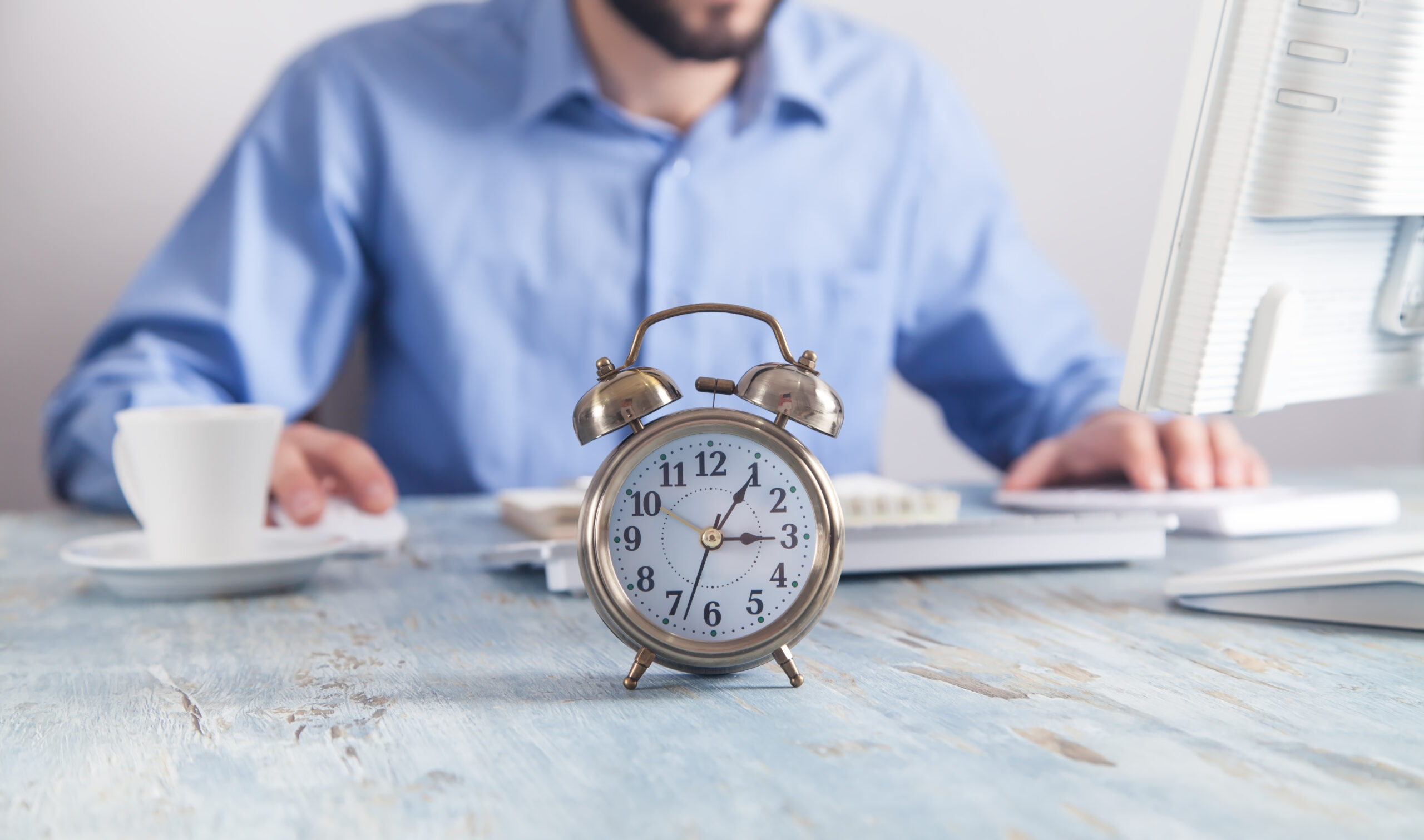 Despertador vintage em primeiro plano sobre mesa de madeira, com homem de camisa azul trabalhando ao computador ao fundo, xícara de café ao lado — representando rotina de trabalho contra o relógio