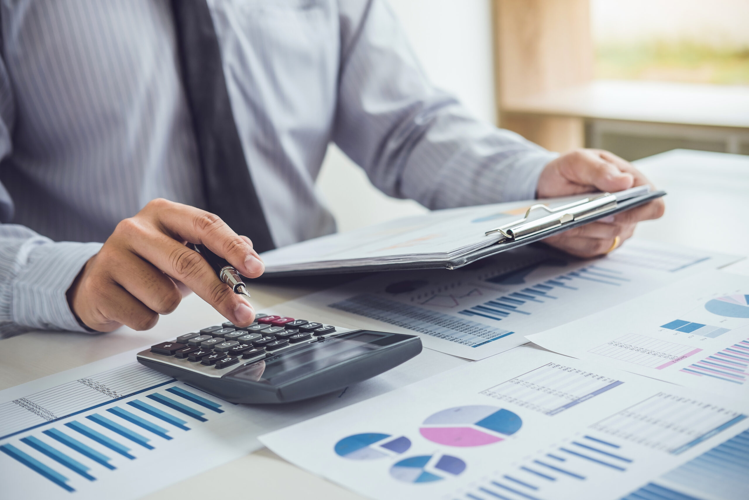 Contador de negócios usando calculadora sobre mesa com diversos relatórios financeiros, gráficos de barras e gráficos de pizza coloridos