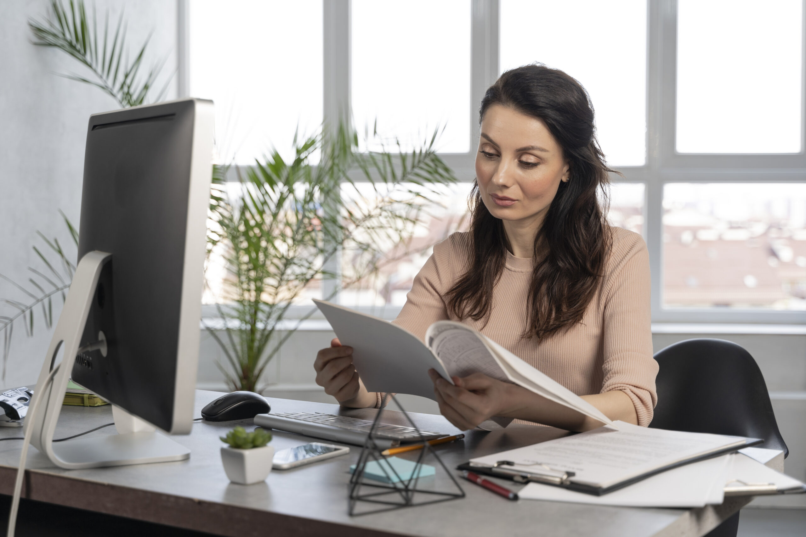 Mulher de cabelos escuros ondulados, vestindo blusa rosé, sentada em uma mesa de escritório lendo documentos, com um monitor de computador, teclado, mouse, celular, vasinho de planta suculenta e prancheta com papéis ao redor; ao fundo, plantas e janelas amplas com vista para a cidade