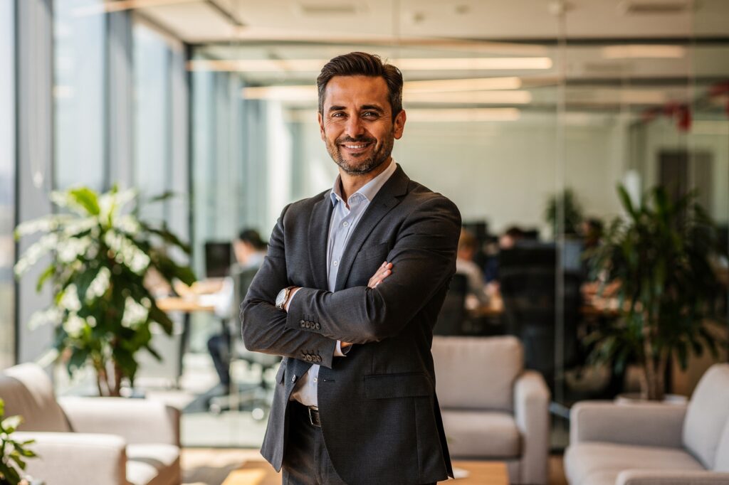 Empresário sorridente de braços cruzados em escritório moderno, com plantas e equipe ao fundo