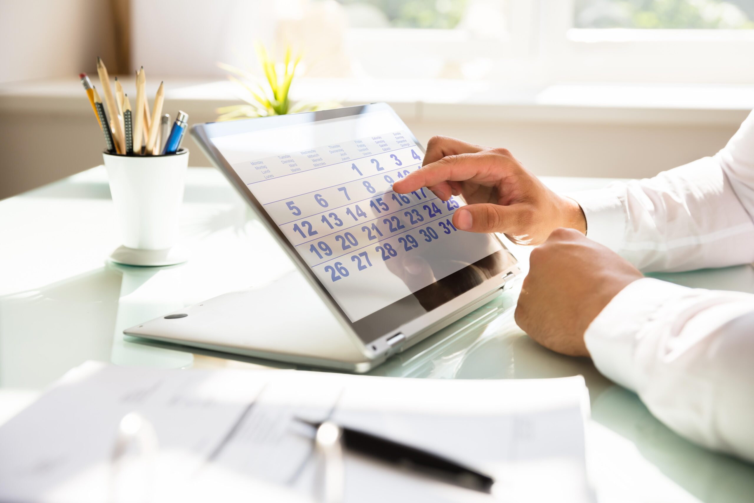 Imagem em close-up de um homem apontando o dedo para um calendário mensal exibido na tela de um laptop conversível (2 em 1), em um ambiente de escritório claro e moderno. O calendário mostra os dias da semana em vários idiomas (inglês, alemão, francês, espanhol) e os números do mês em azul. Ao fundo, veem-se um porta-canetas com lápis, uma planta desfocada e uma janela iluminada. Documentos em papel estão apoiados na mesa de vidro em primeiro plano.
