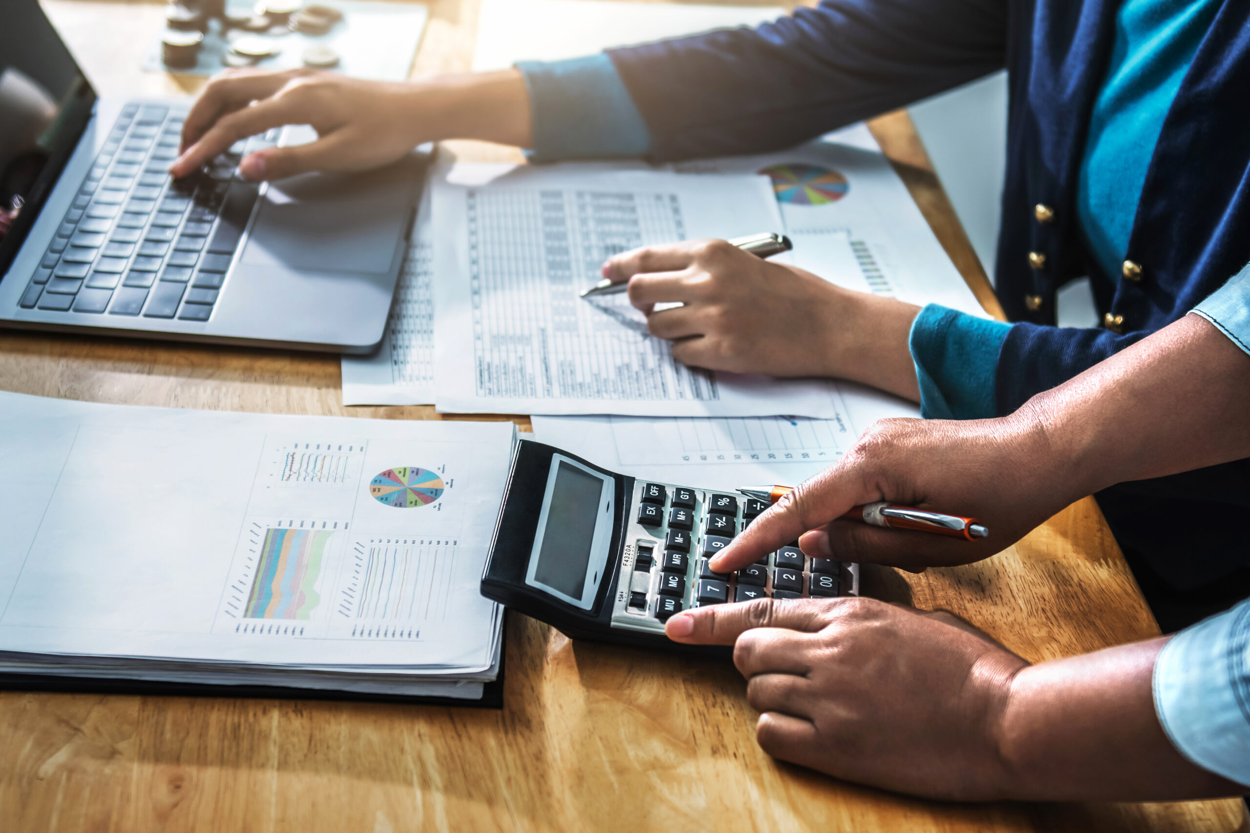 Duas pessoas analisando relatórios financeiros com gráficos, usando calculadora e laptop durante reunião de auditoria contábil.