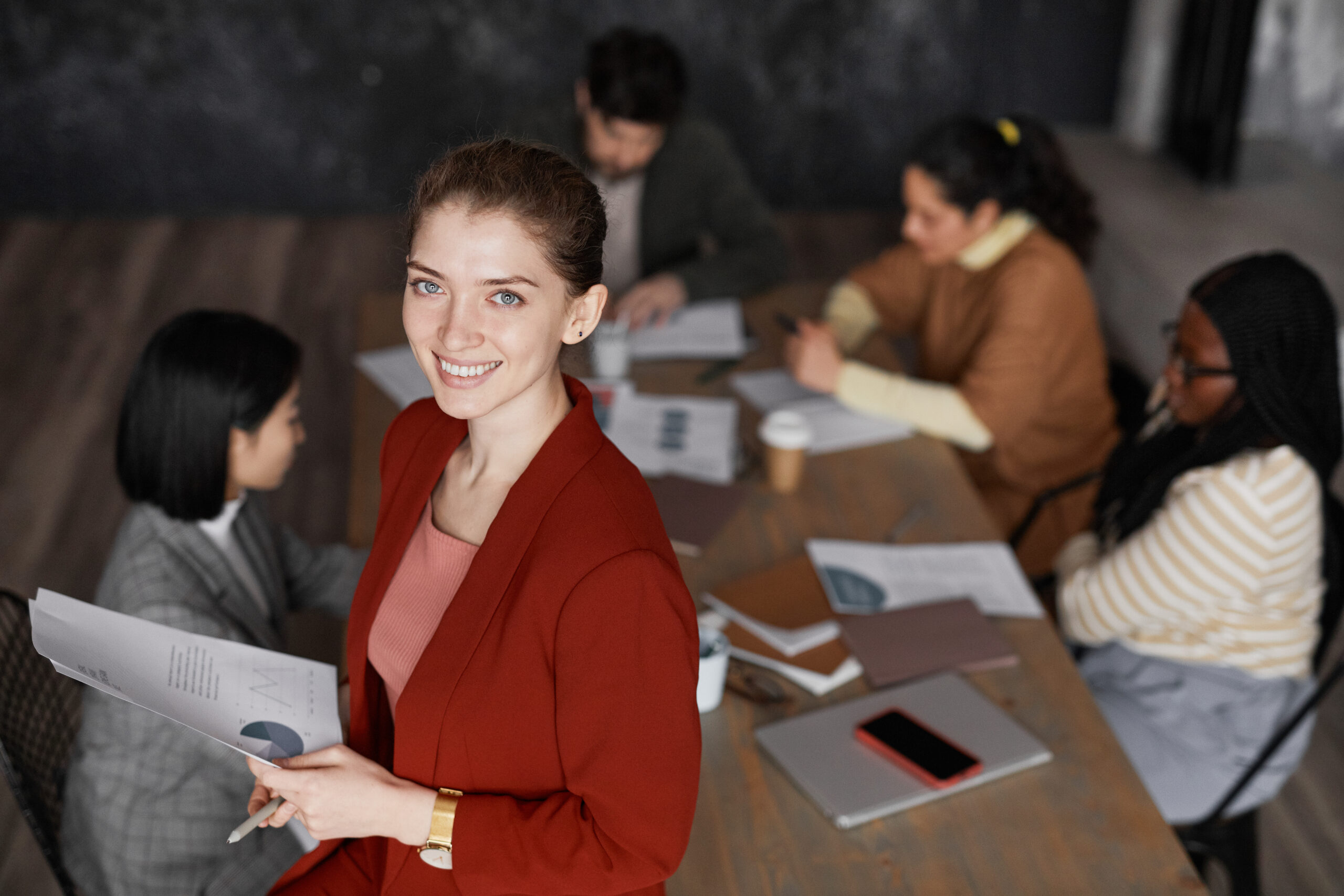 Empresária sorridente de blazer vermelho segurando relatório com gráficos, em primeiro plano, com equipe reunida ao fundo em mesa de reunião com laptop, celular e documentos.