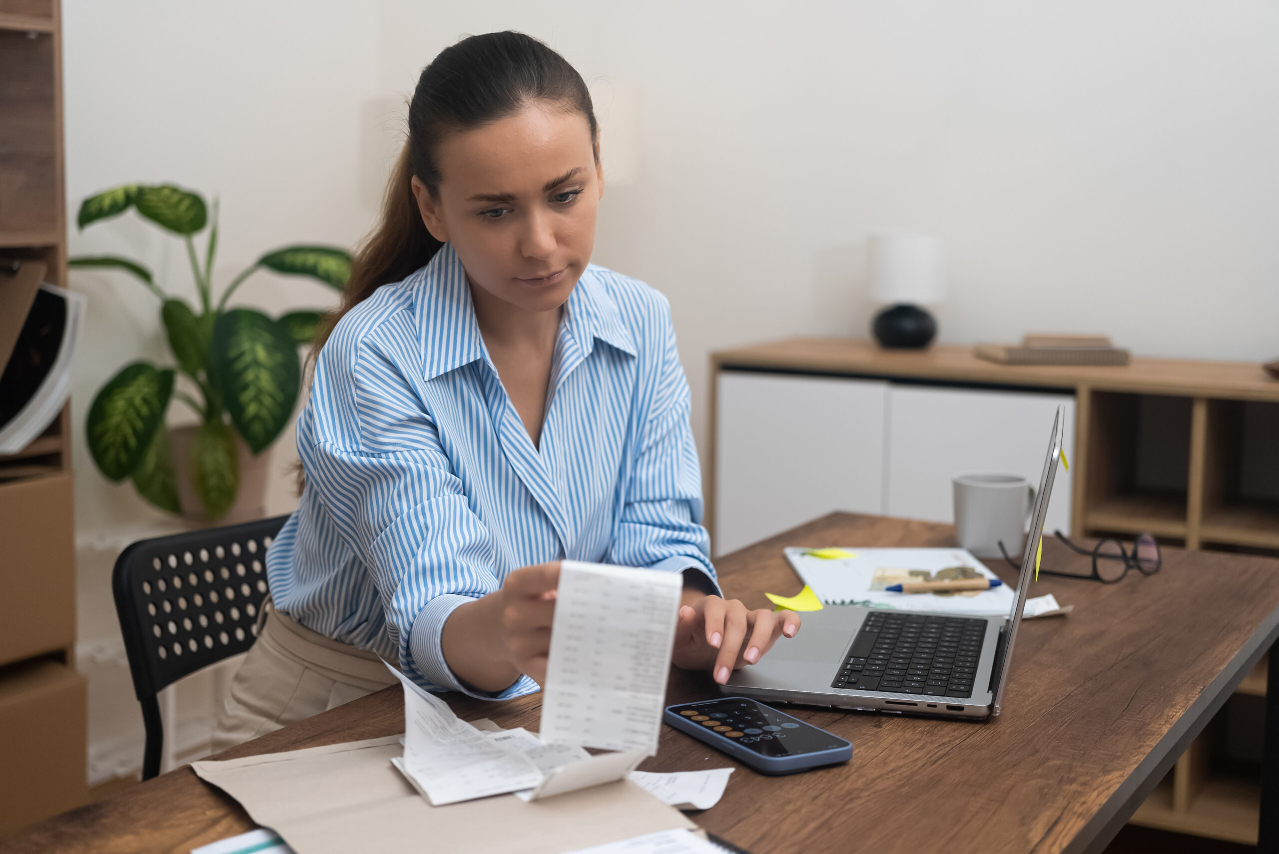 Mulher analisando contas e documentos financeiros em casa com notebook e calculadora.