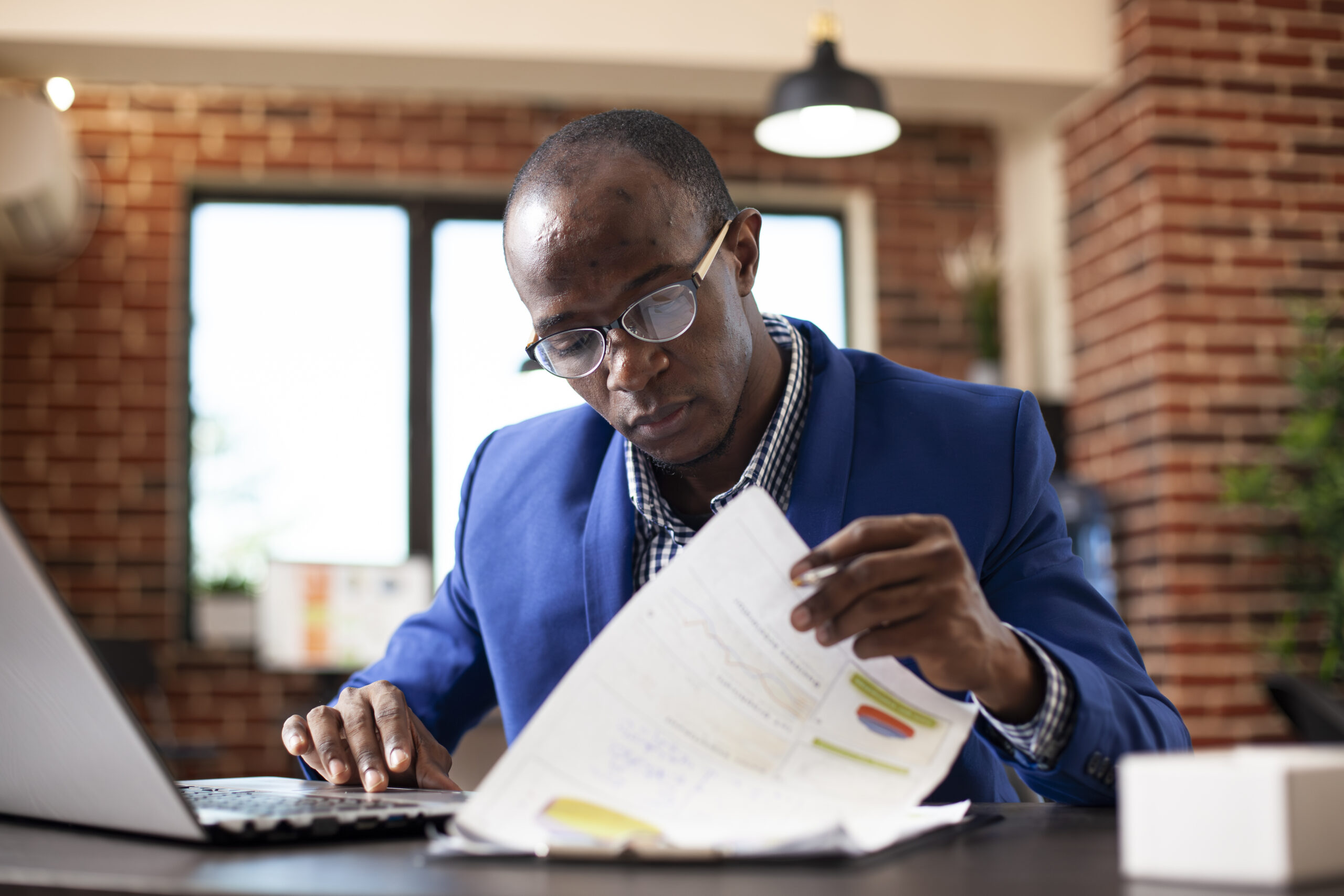 Homem de terno azul e óculos analisando um relatório impresso com gráficos coloridos enquanto digita em um notebook, em um ambiente de escritório com parede de tijolos aparentes