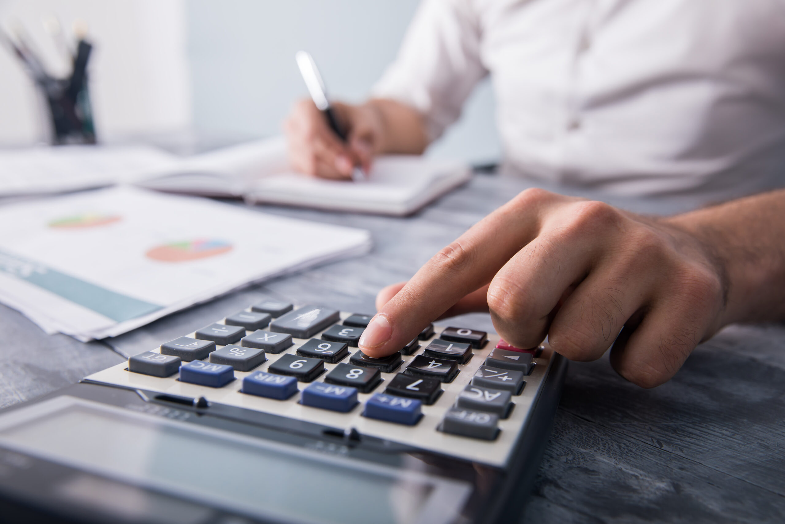 Homem usando calculadora e anotando dados financeiros em caderno, com relatório de gráficos sobre a mesa