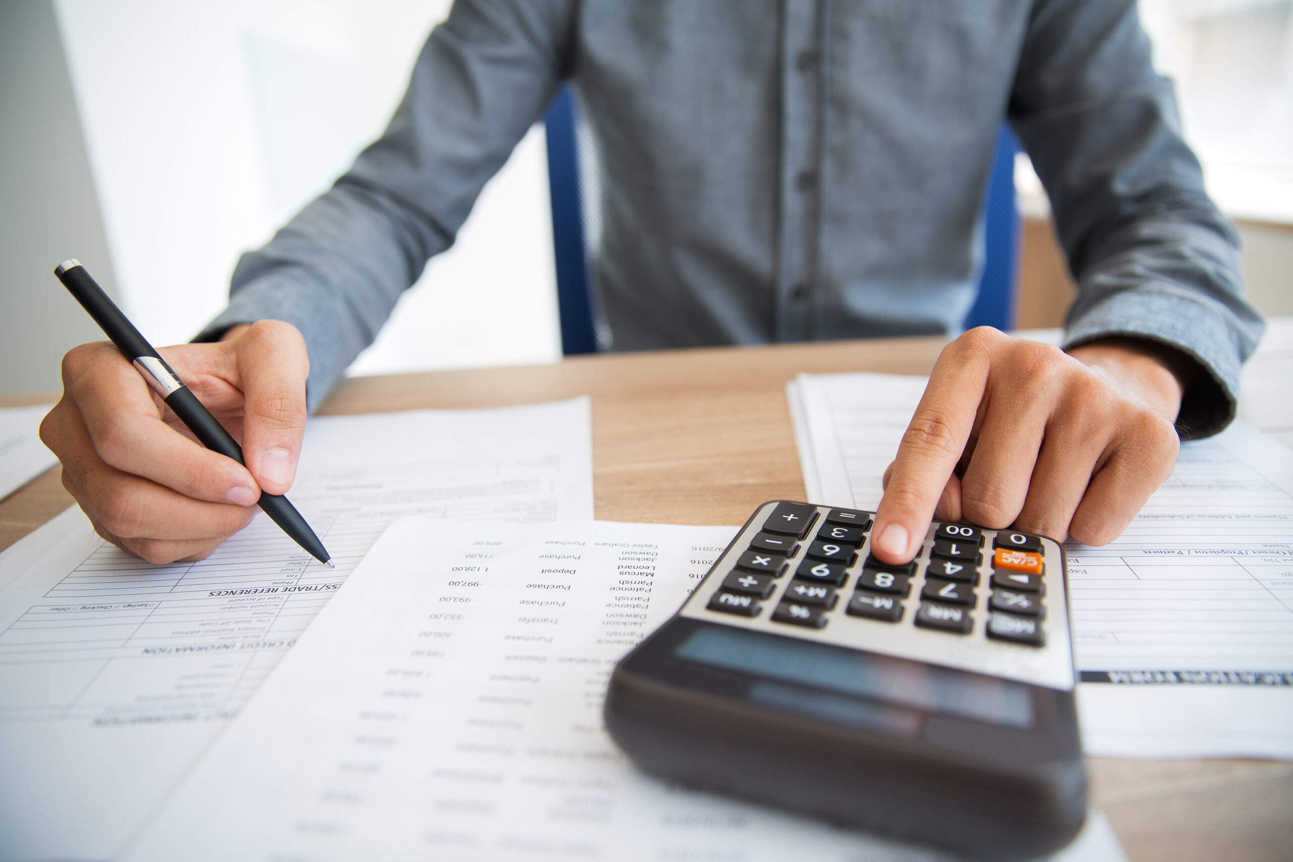 Close-up de um homem com camisa social cinza usando uma calculadora enquanto segura uma caneta, com documentos financeiros — incluindo um extrato com referências comerciais e uma planilha de transações — espalhados sobre a mesa de madeira