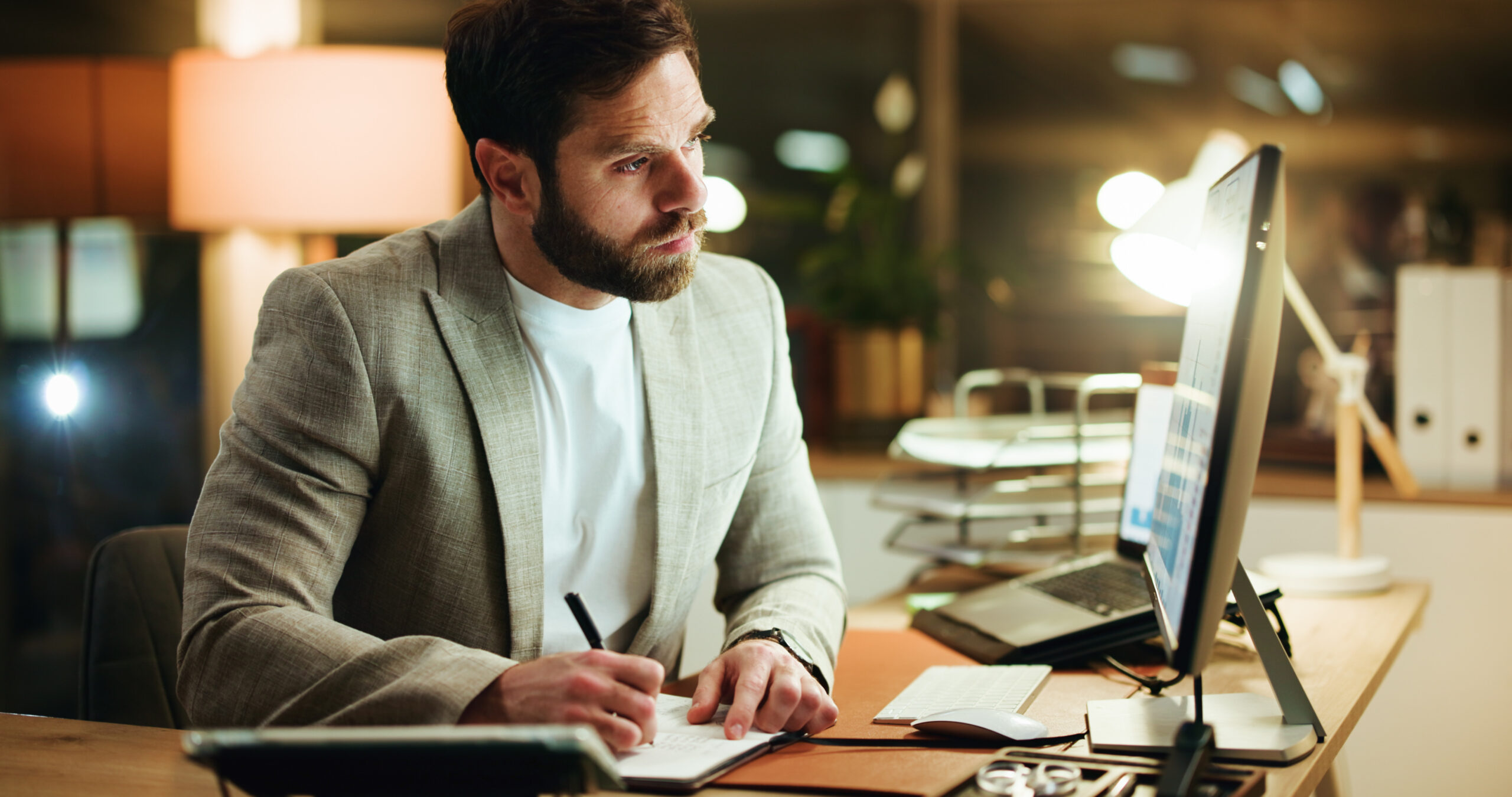 um homem de barba, vestindo blazer sobre camiseta branca, sentado à mesa à noite, escrevendo em um caderno enquanto olha para um monitor com dados/gráficos na tela. O ambiente é um escritório com iluminação quente (luminária ao fundo), teclado, mouse, laptop e óculos sobre a mesa.