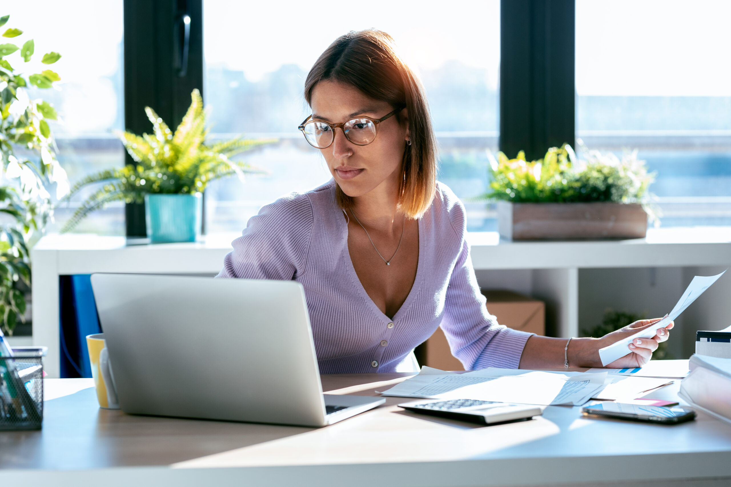 Mulher jovem concentrada trabalhando em um laptop no escritório em casa, usando óculos, enquanto analisa documentos financeiros com uma calculadora e papéis sobre a mesa, com plantas e janela ao fundo.