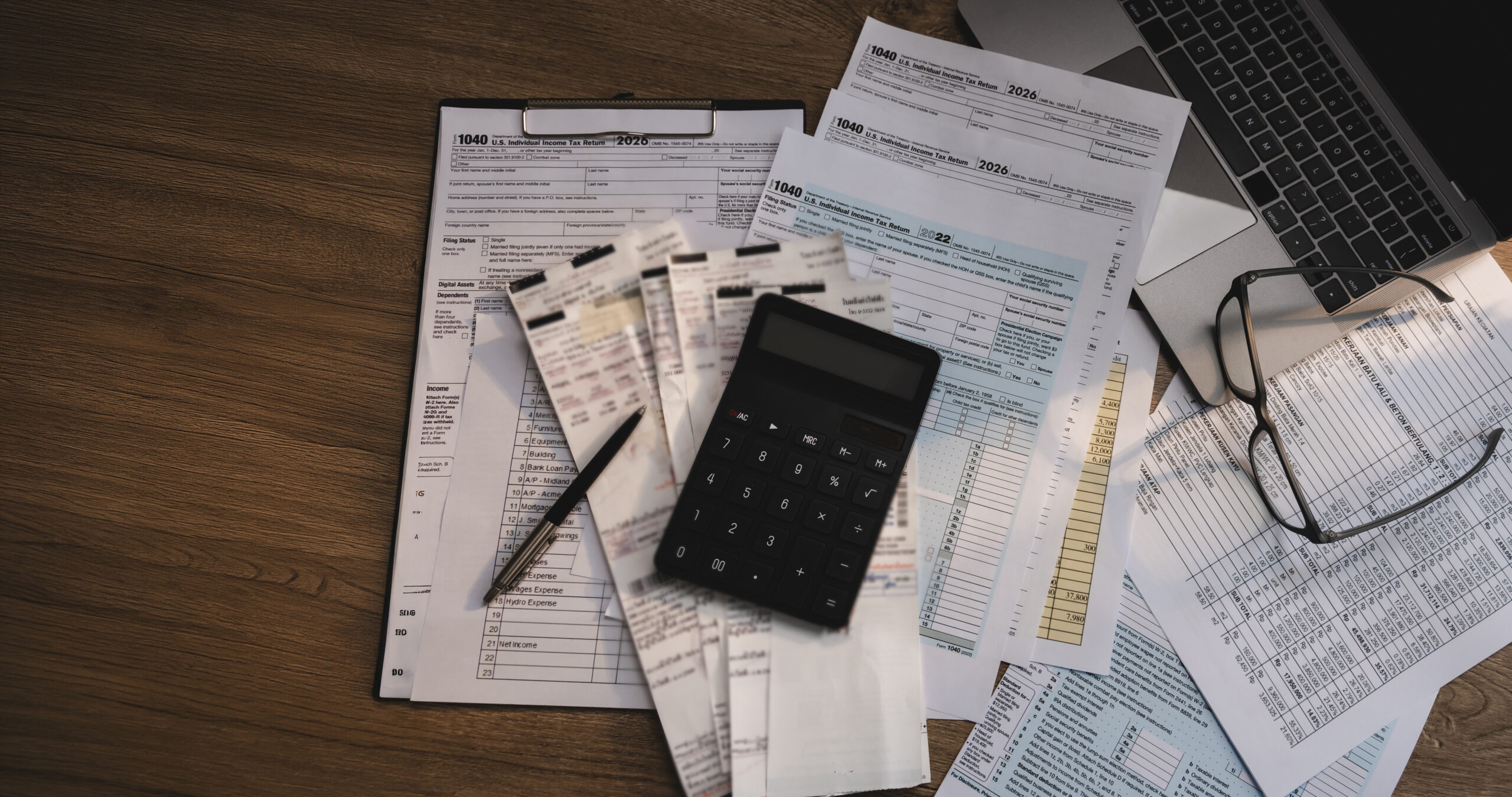 "Mesa de madeira com formulários de imposto de renda (Form 1040), recibos, calculadora, caneta, óculos e notebook ao fundo — cenário de preparação e revisão fiscal.