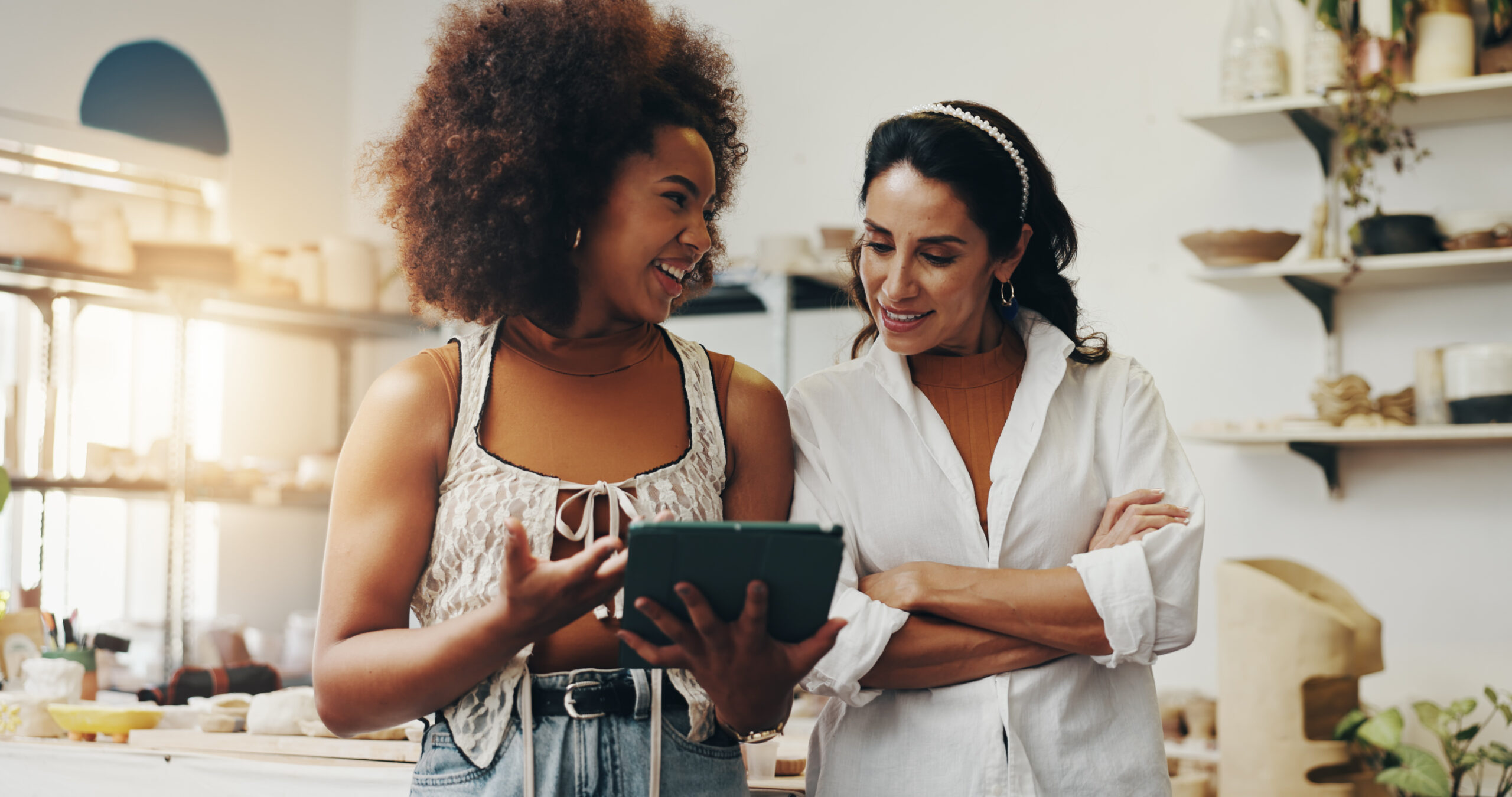 Duas mulheres em um ateliê de cerâmica, sorrindo e olhando juntas para um tablet. Uma delas usa um top de renda branca e segura o tablet, gesticulando enquanto fala; a outra, de camisa branca e faixa de pérolas no cabelo, está com os braços cruzados enquanto acompanha a tela. Ao fundo, prateleiras com peças de cerâmica, potes e plantas, com luz solar entrando pela janela à esquerda.