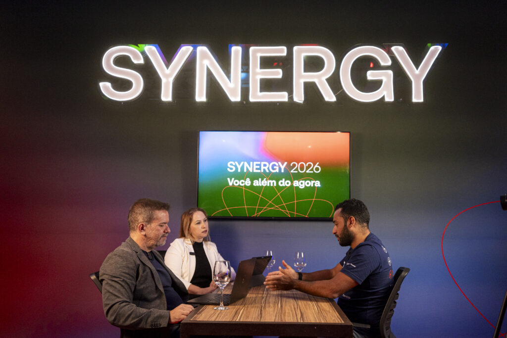Três pessoas conversam em uma mesa de madeira com notebook e taças, em um estúdio do Synergy 2026. Um homem de camiseta azul com a marca Domínio gesticula enquanto explica algo a um homem de blazer cinza e a uma mulher de blazer branco. Ao fundo, o letreiro neon "SYNERGY" e uma TV com a frase "Synergy 2026 – Você além do agora