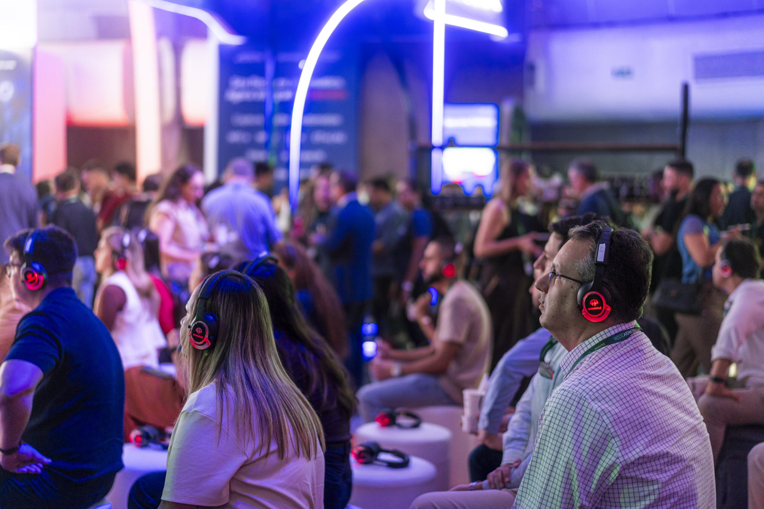 Público do evento Synergy 2026 sentado em pufes, usando fones de ouvido com luz vermelha enquanto assiste a uma palestra. Ao fundo, participantes circulam pelo espaço, iluminado com luzes neon em tons de azul e roxo.