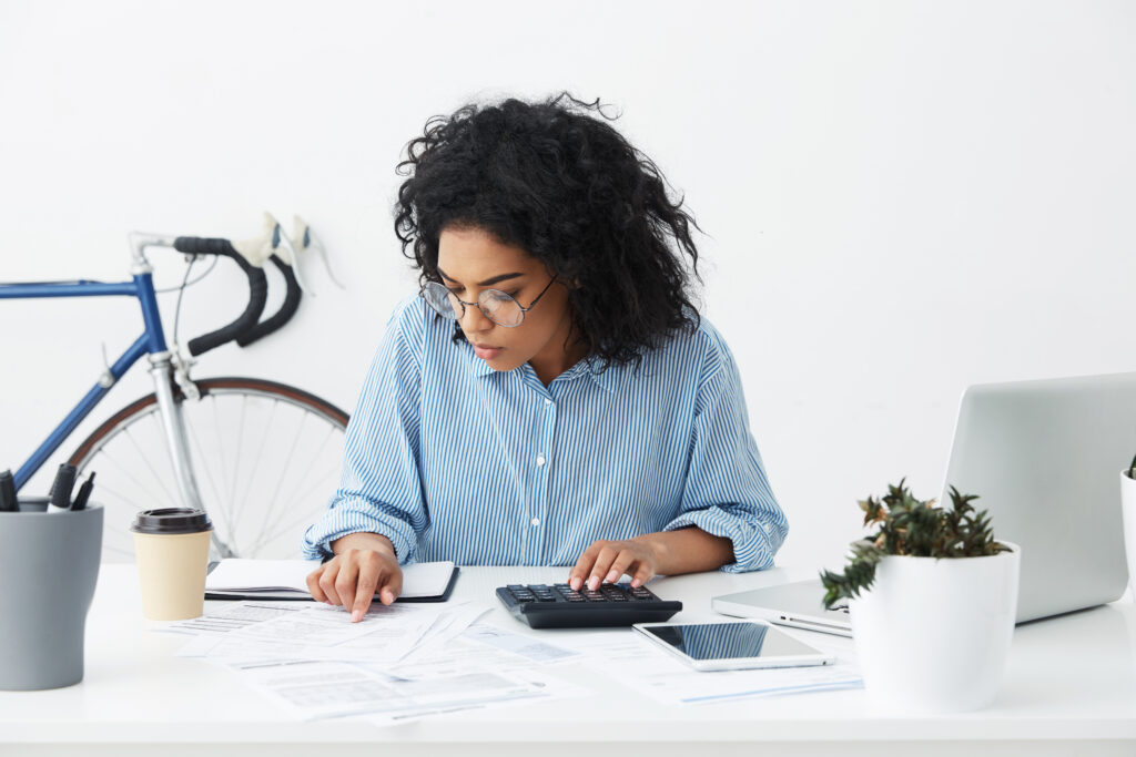 Profissional de óculos confere documentos impressos e usa calculadora em mesa de escritório, com notebook, tablet e copo de café.