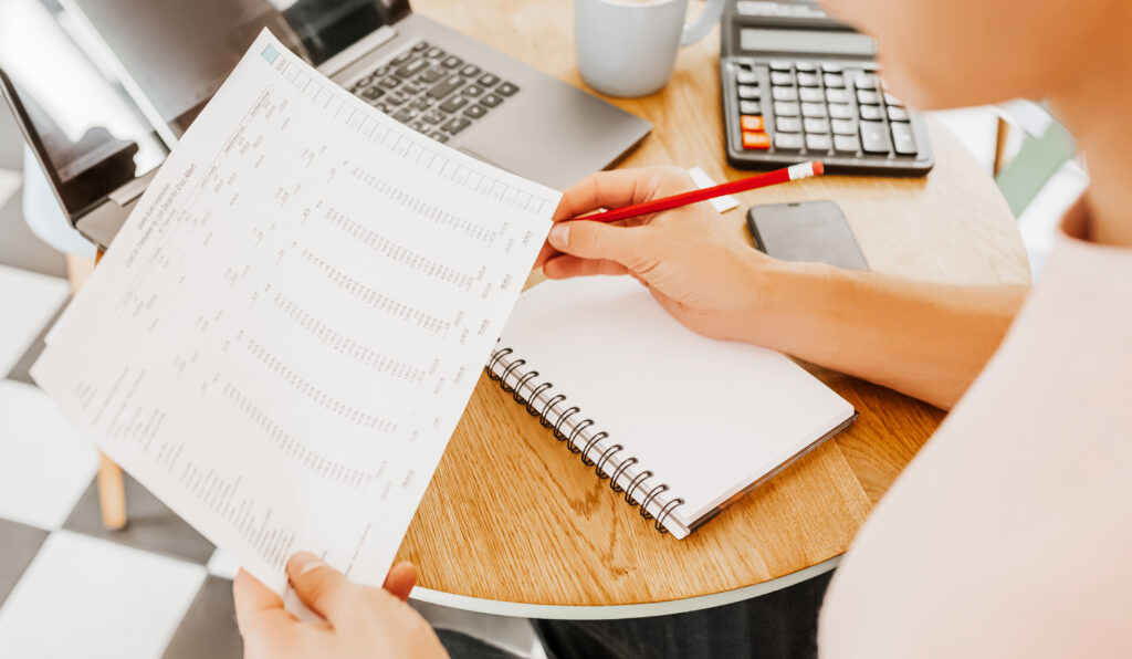 Pessoa analisa relatório financeiro impresso com lápis vermelho na mão, sobre mesa com caderno, calculadora, notebook e celular.