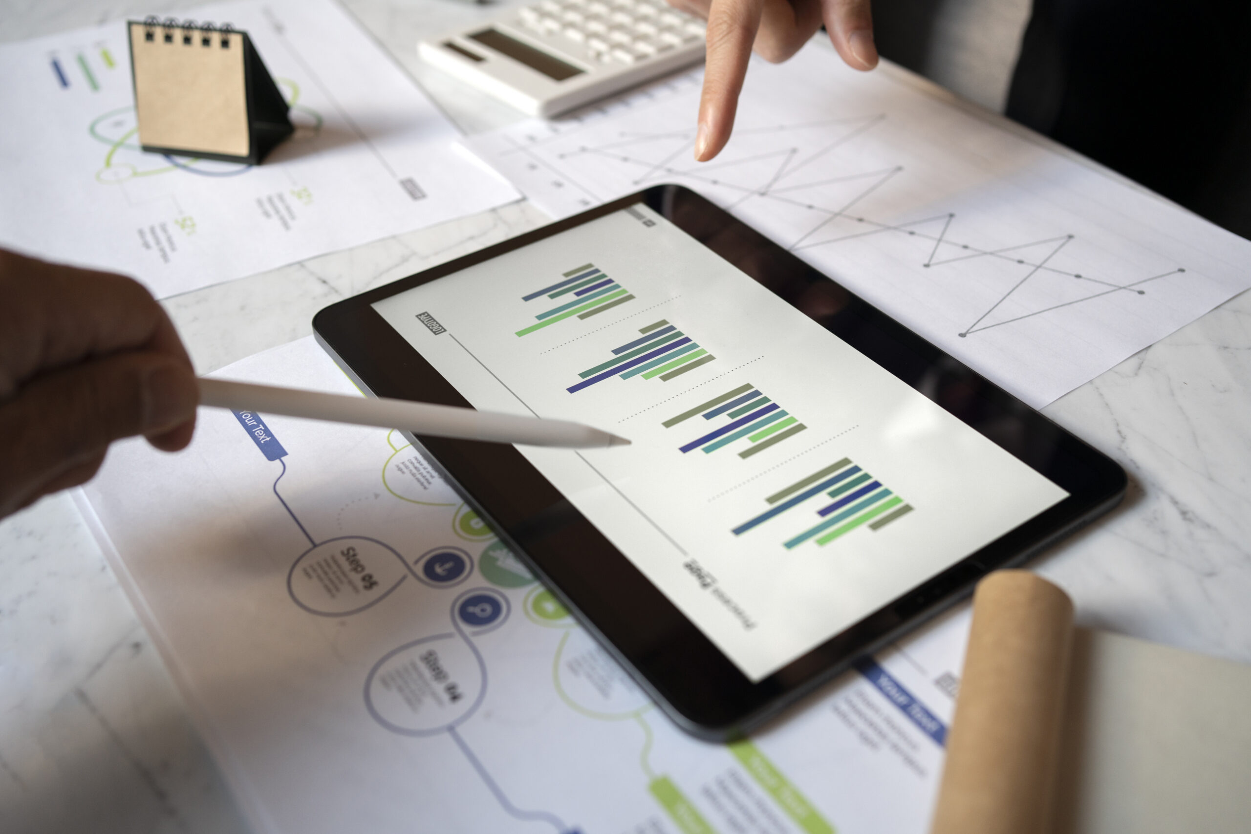 Contadores analisam gráficos de barras em um tablet, com relatórios impressos, calculadora e calendário sobre a mesa