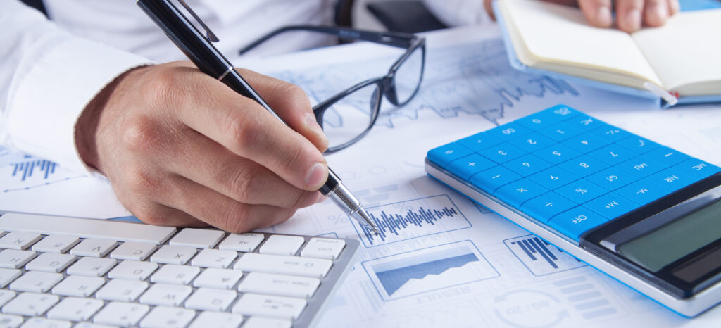 Mão preenchendo gráficos financeiros com uma caneta sobre documentos com indicadores e relatórios, ao lado de uma calculadora azul, teclado de computador e óculos, representando análise financeira e gestão contábil.