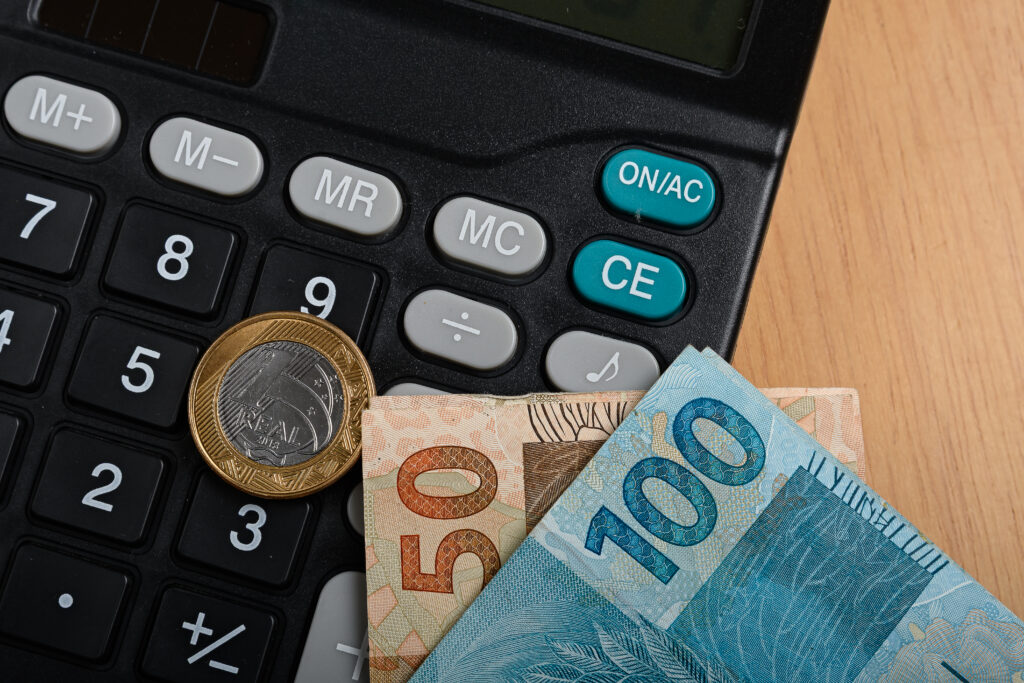 Calculadora preta com moeda de 1 real e notas de 50 e 100 reais sobre uma mesa de madeira.