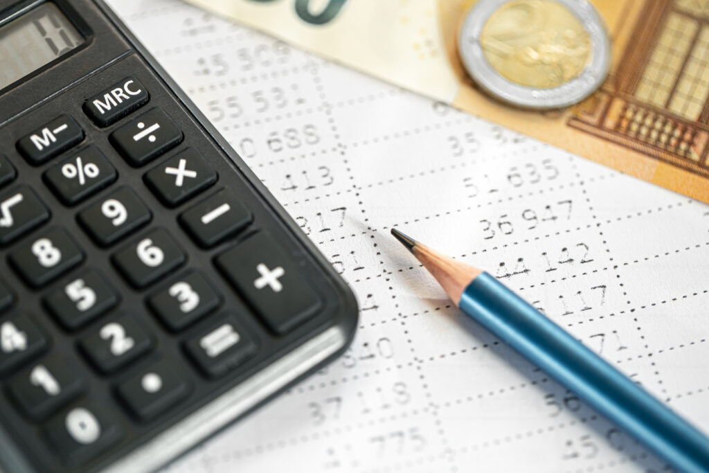 Calculadora preta ao lado de um lápis azul sobre uma planilha impressa com números, com notas e moeda de euro ao fundo.