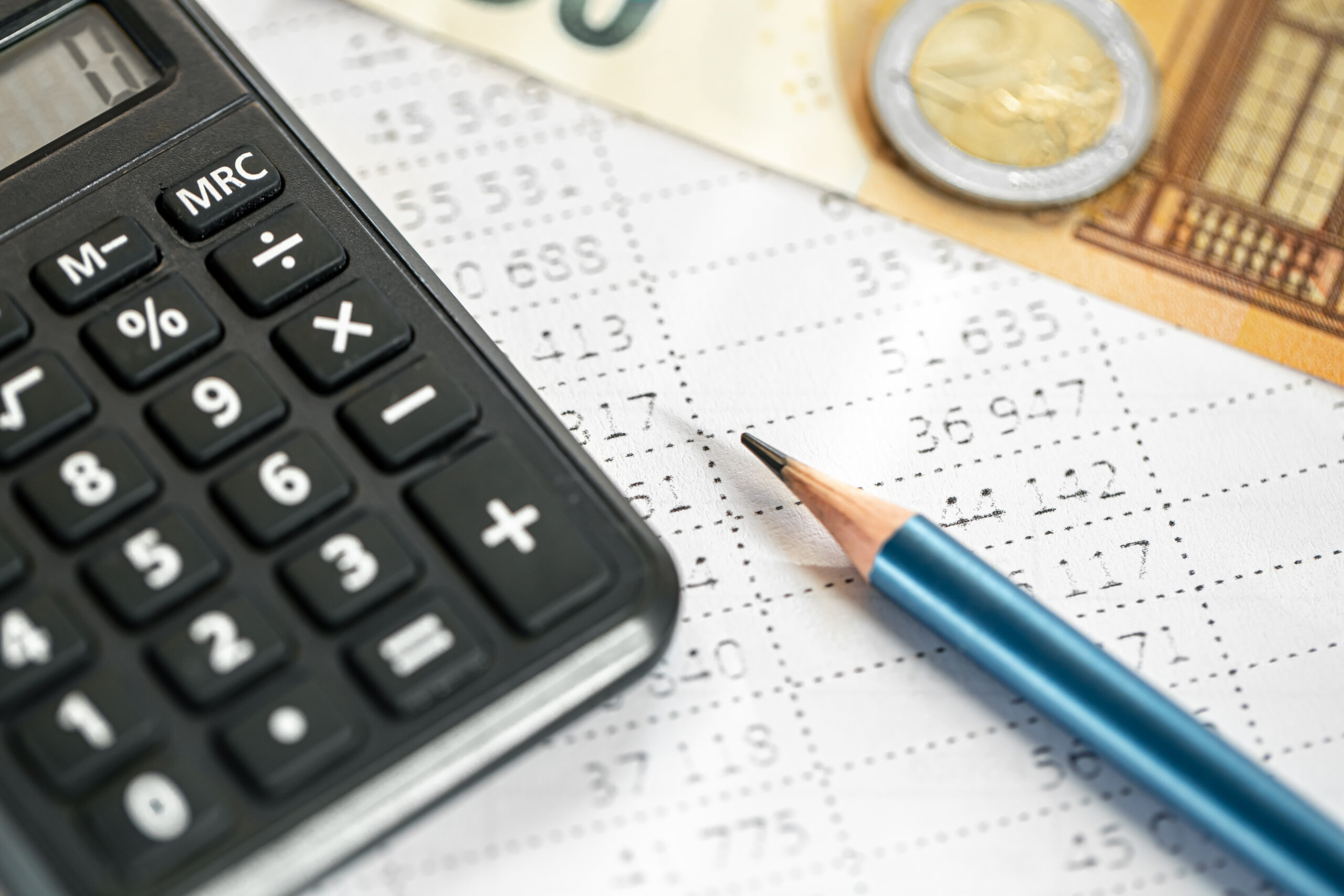 Calculadora preta ao lado de um lápis azul sobre uma planilha impressa com números, com notas e moeda de euro ao fundo.