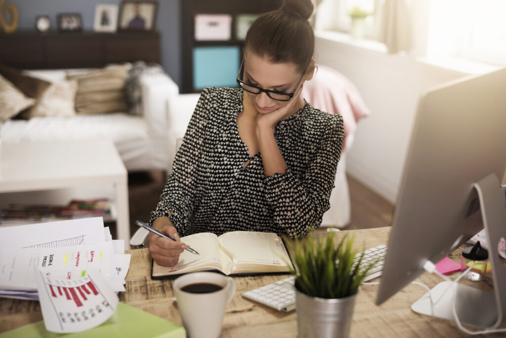 Mulher de óculos fazendo anotações em uma agenda em seu home office, com computador, café e gráficos impressos sobre a mesa