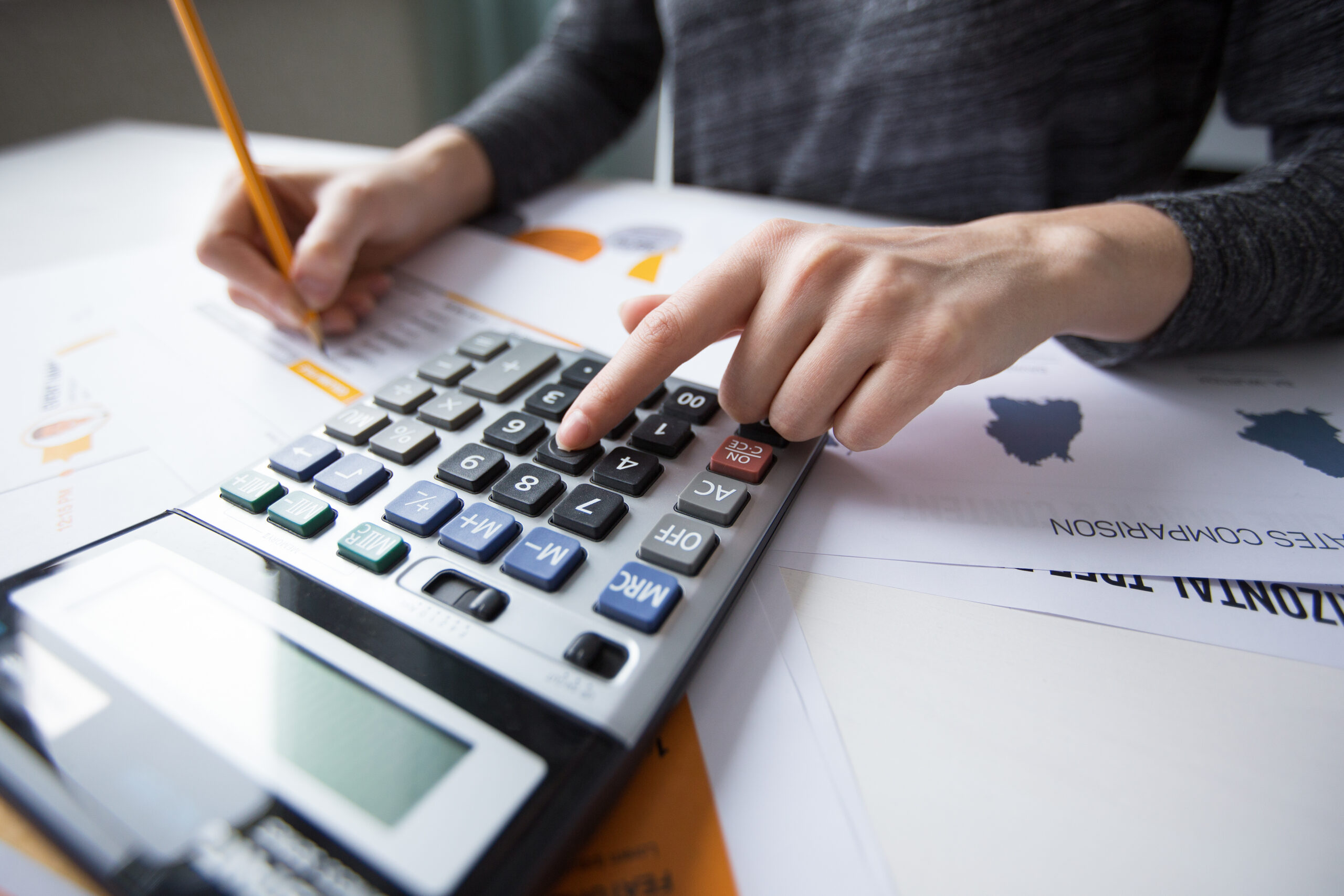 Profissional usando calculadora e fazendo anotações a lápis sobre relatórios impressos com gráficos.