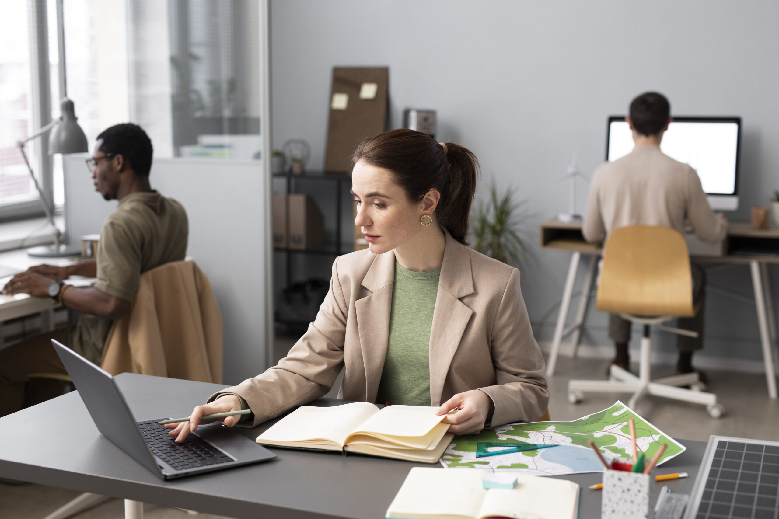 Profissional analisa anotações e trabalha no notebook em escritório compartilhado, com colegas em suas estações ao fundo.