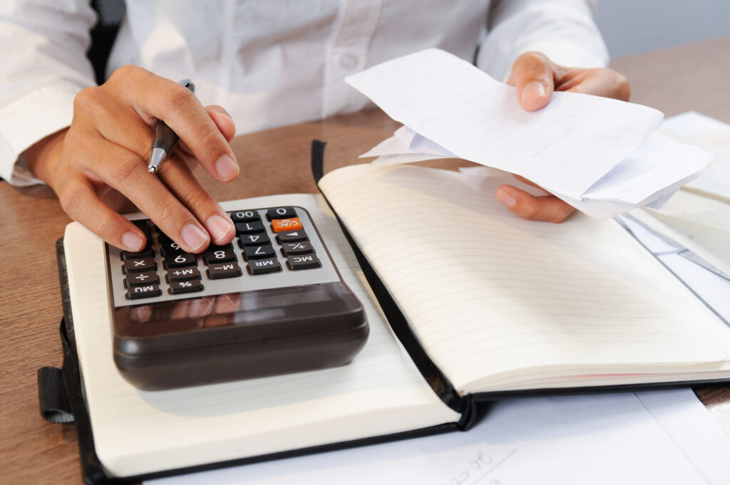 Pessoa de camisa branca usando calculadora sobre um caderno aberto enquanto segura comprovantes de compra.
