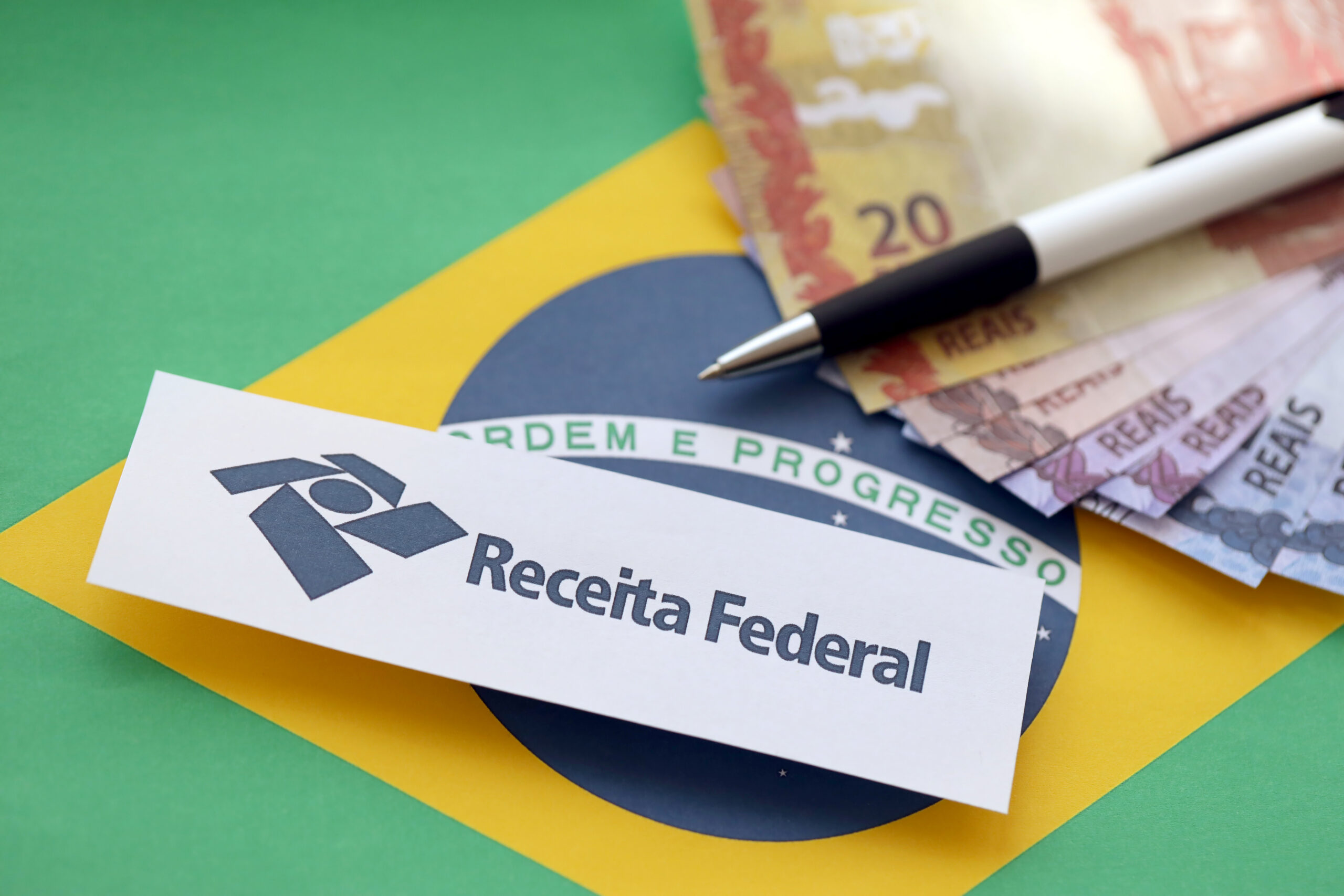 Papel com o logotipo e o nome da Receita Federal sobre a bandeira do Brasil, ao lado de cédulas de real e uma caneta.