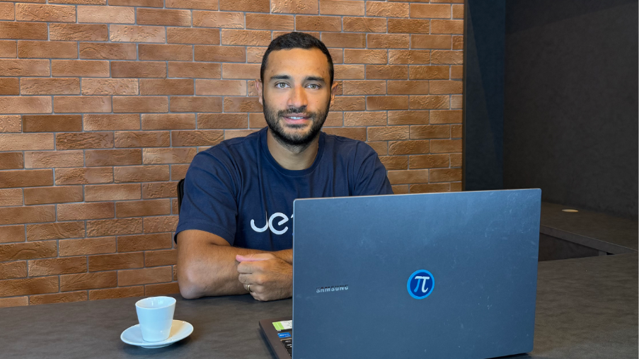 Gustavo Dacol, CEO da Jettax, de camiseta azul-marinho com a marca da Jettax, sentado à mesa com notebook e xícara de café, em escritório com parede de tijolos aparentes.
