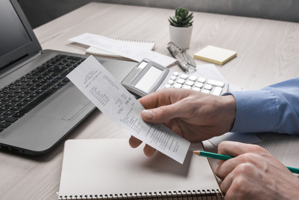 Pessoa analisando um comprovante com calculadora, notebook e caderno sobre a mesa, representando o controle financeiro e a apuração de impostos.