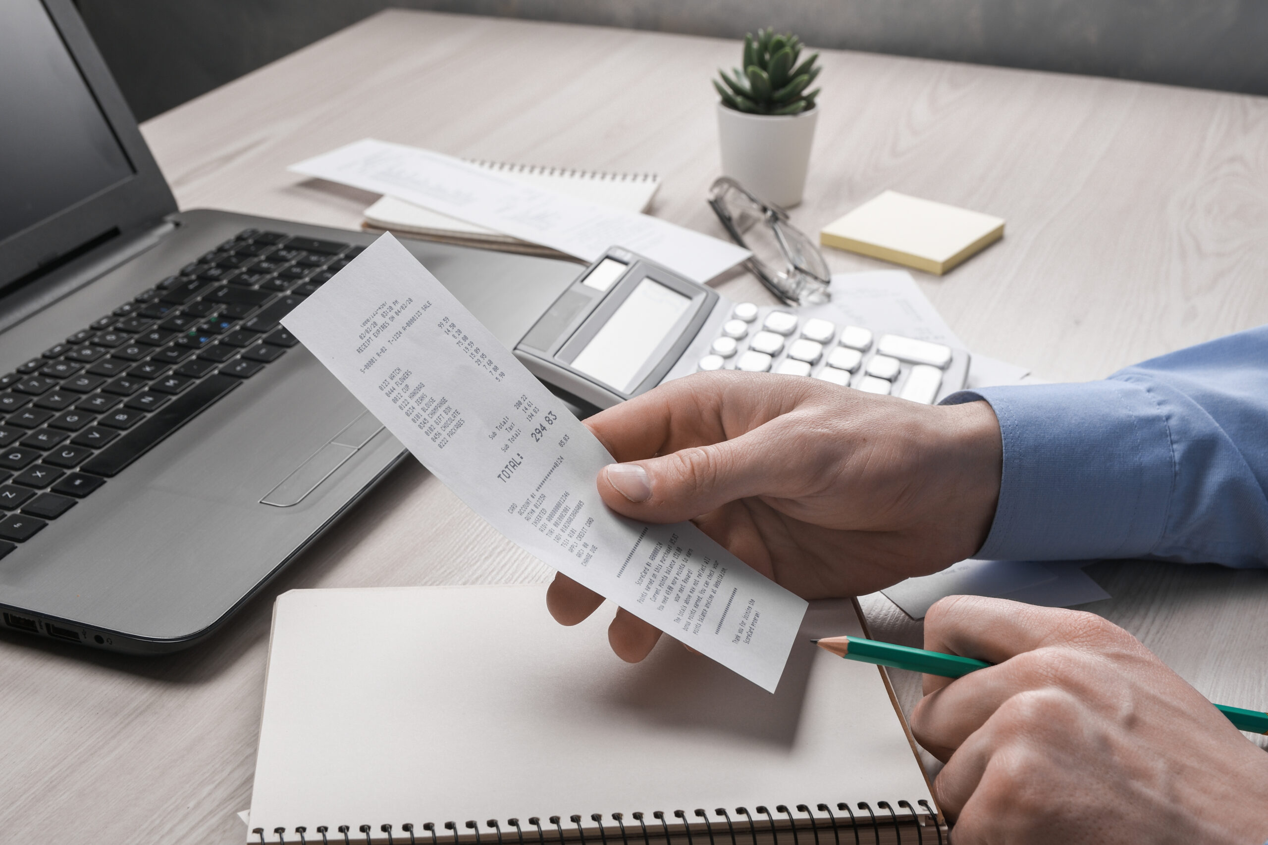 Pessoa analisando um comprovante com calculadora, notebook e caderno sobre a mesa, representando o controle financeiro e a apuração de impostos.