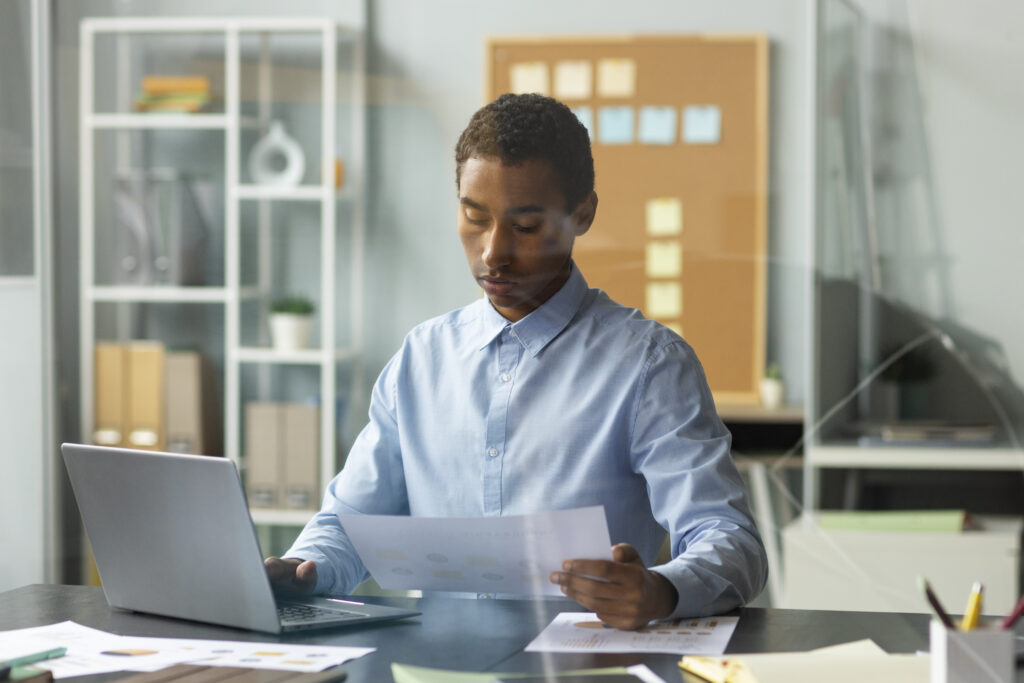Profissional de camisa azul analisa relatório impresso com gráficos enquanto usa um notebook em mesa de escritório