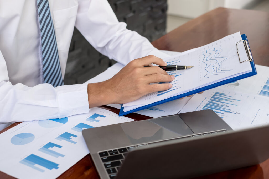 Profissional de camisa social e gravata analisa relatórios financeiros com gráficos de barras e linhas em uma prancheta, ao lado de um notebook sobre a mesa.