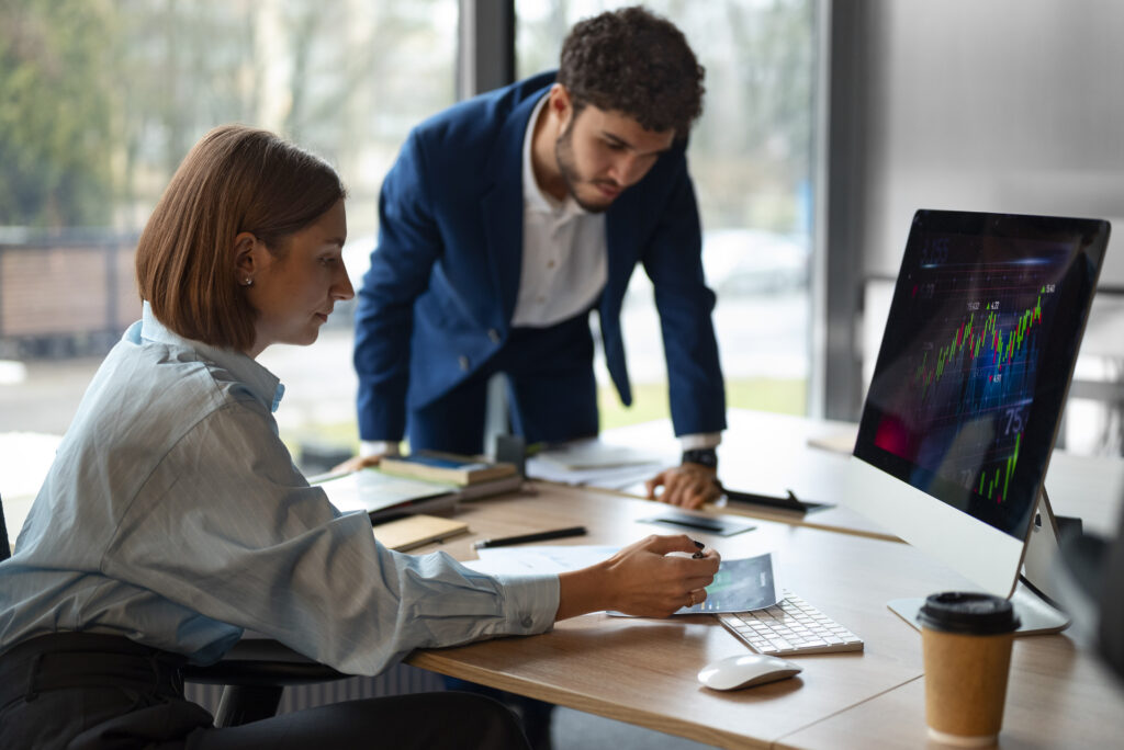 Profissional analisa gráfico impresso sobre a mesa enquanto um colega de terno observa, ao lado de um monitor com gráficos financeiros.