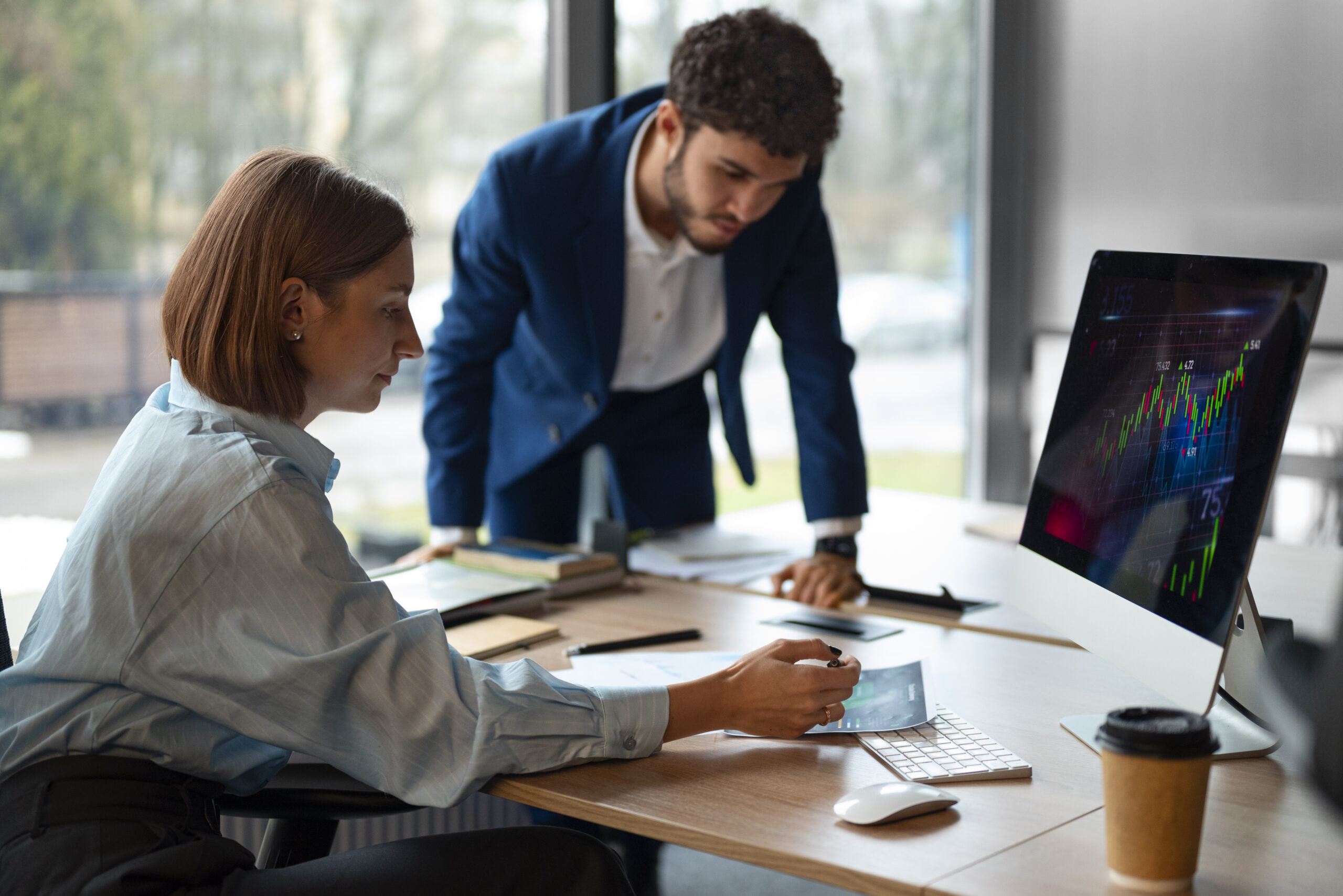 Profissional analisa gráfico impresso sobre a mesa enquanto um colega de terno observa, ao lado de um monitor com gráficos financeiros.