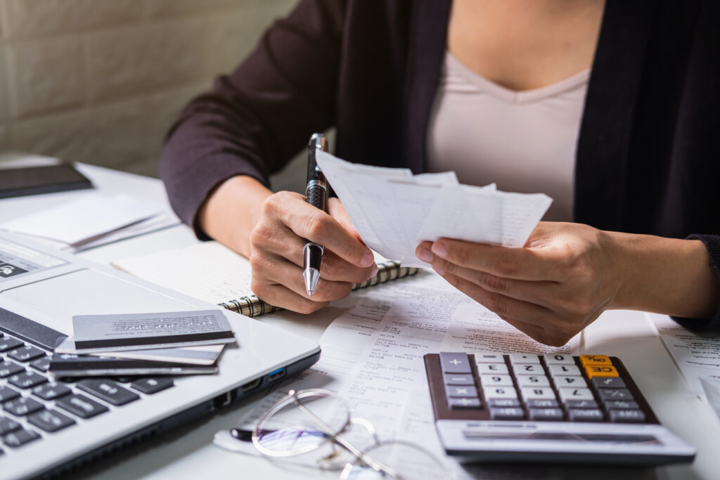 Mulher analisando notas fiscais e recibos com caneta na mão, em mesa com notebook, cartões de crédito, calculadora e documentos financeiros.