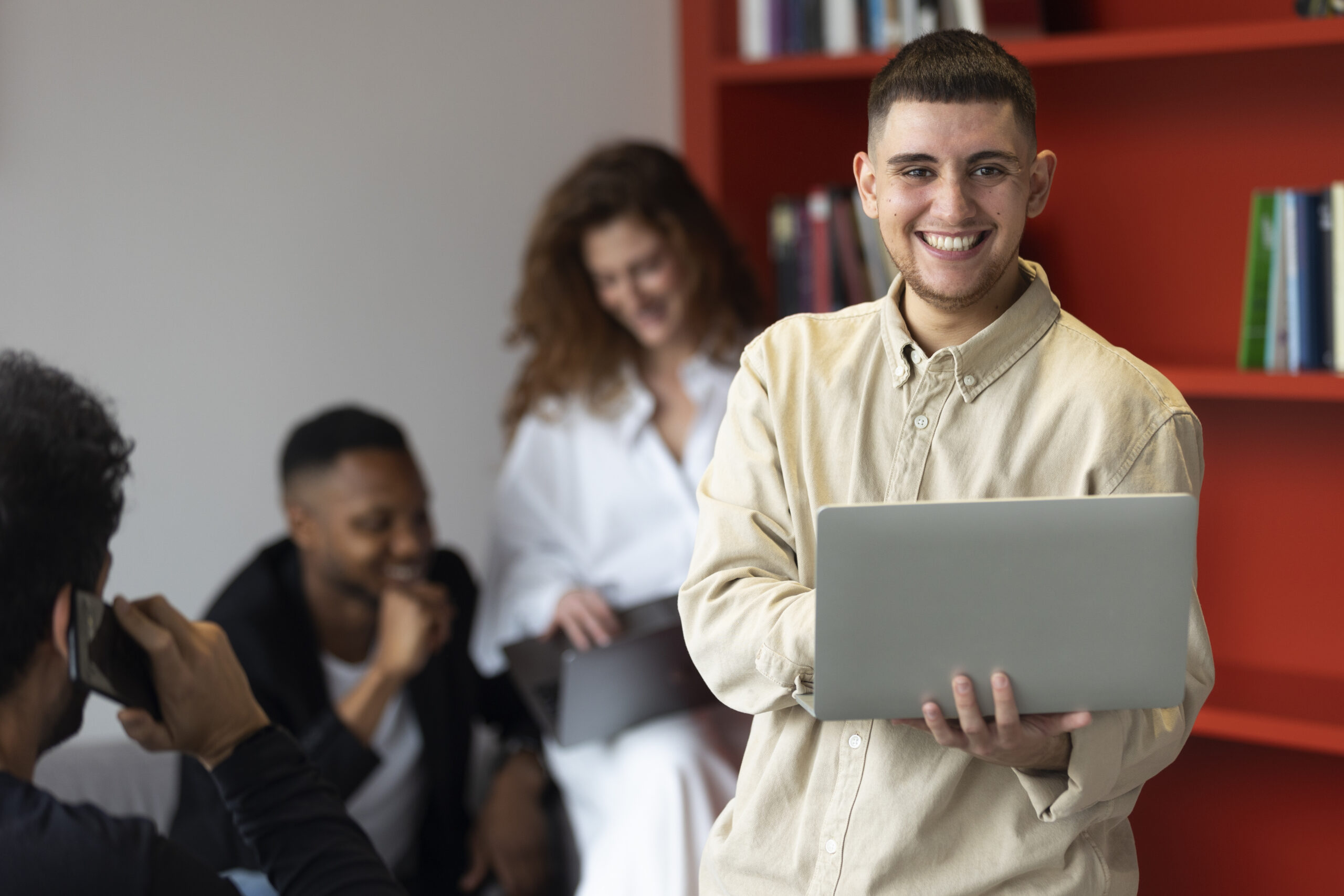 Homem sorridente segurando um notebook em um escritório, em frente a uma estante vermelha com livros. Ao fundo, colegas conversam e usam notebook e celular.