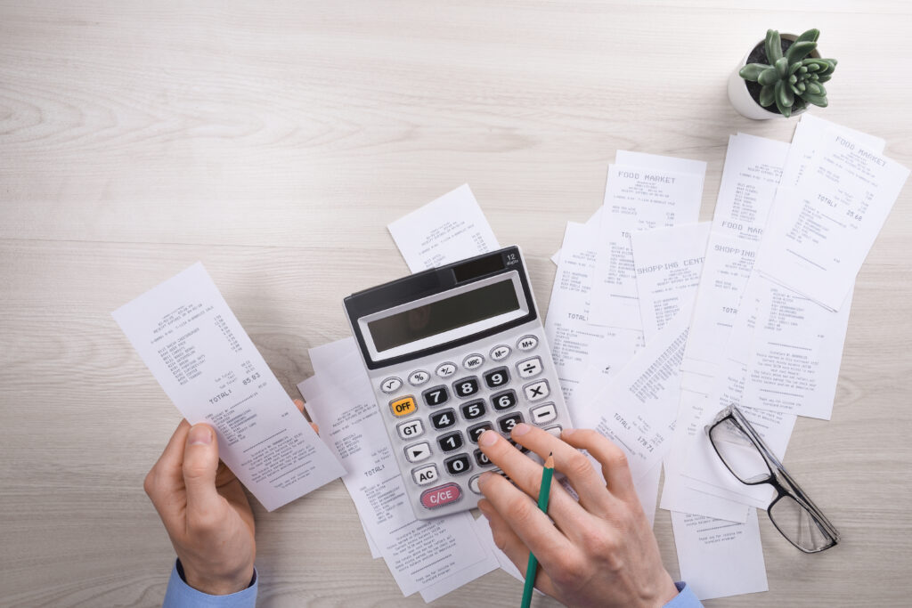 Mãos segurando um cupom fiscal e usando calculadora sobre uma mesa com diversos recibos, óculos e um vaso de planta.