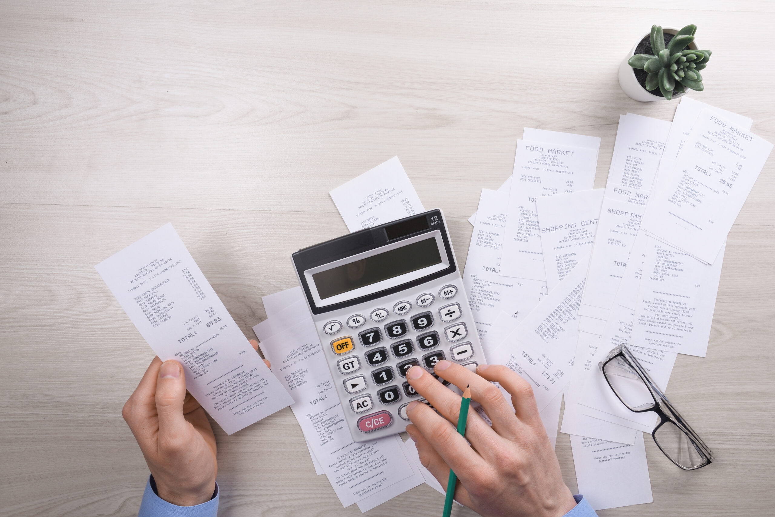 Mãos segurando um cupom fiscal e usando calculadora sobre uma mesa com diversos recibos, óculos e um vaso de planta.