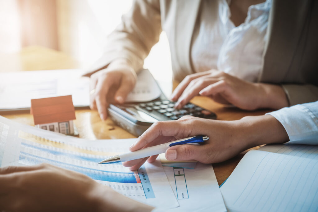 Profissionais analisam relatório financeiro com caneta e calculadora sobre a mesa, ao lado de uma miniatura de casa.