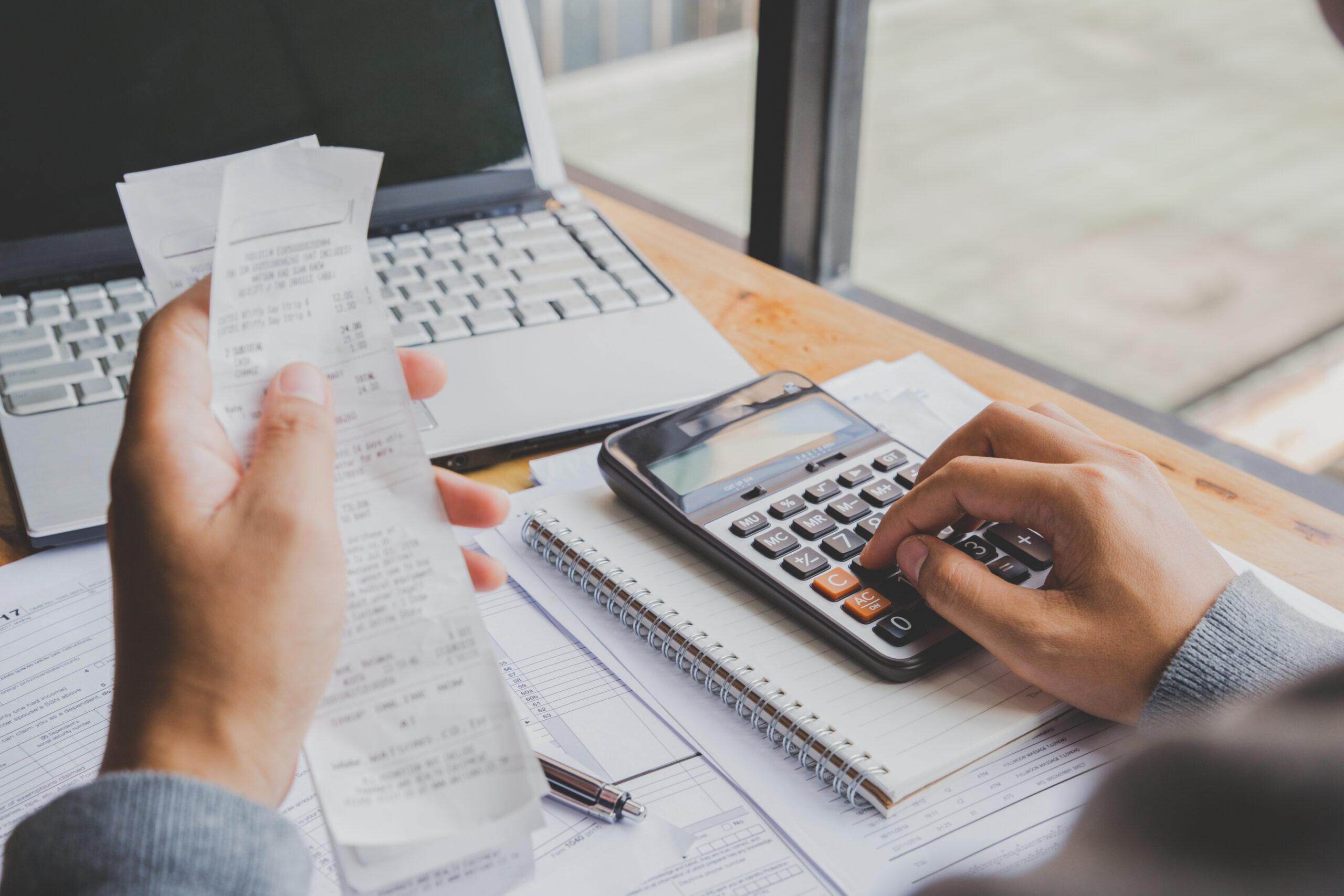 Profissional de contabilidade confere cupons fiscais e digita na calculadora, com notebook e documentos sobre a mesa.